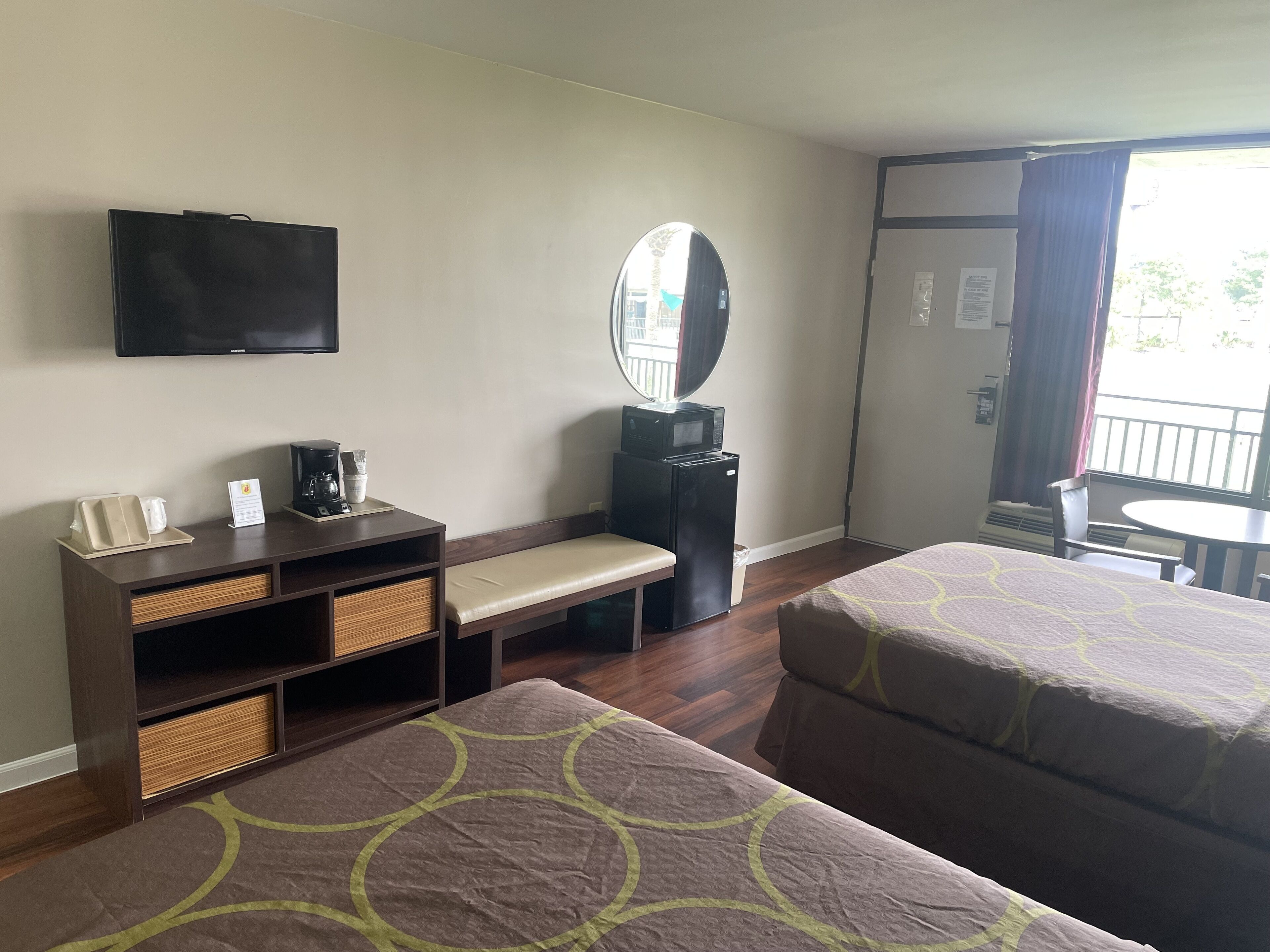 Ein Hotelzimmer mit zwei Einzelbetten, einem Schreibtisch mit Kaffeemaschine, einem Flachbildfernseher, einem Spiegel und einem Fenster mit Vorhängen.