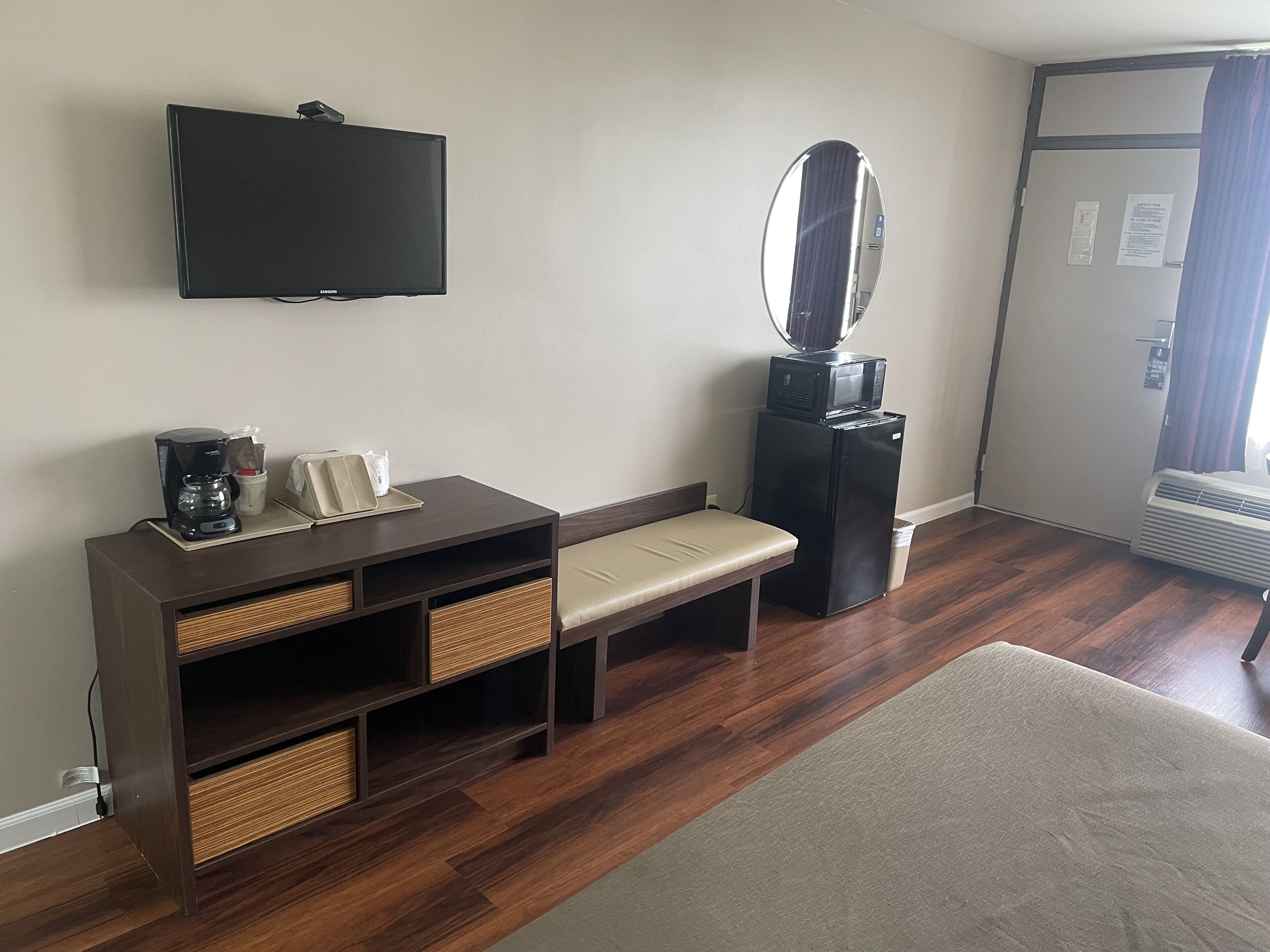 A hotel room with a flat-screen TV, a small table with a coffee maker, a bench, a mirror, and a door with a window.