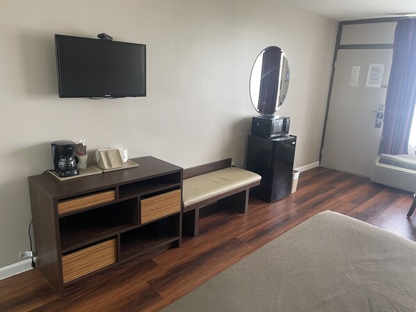 A hotel room with a flat-screen TV, a small table with a coffee maker, a bench, a mirror, and a door with a window.