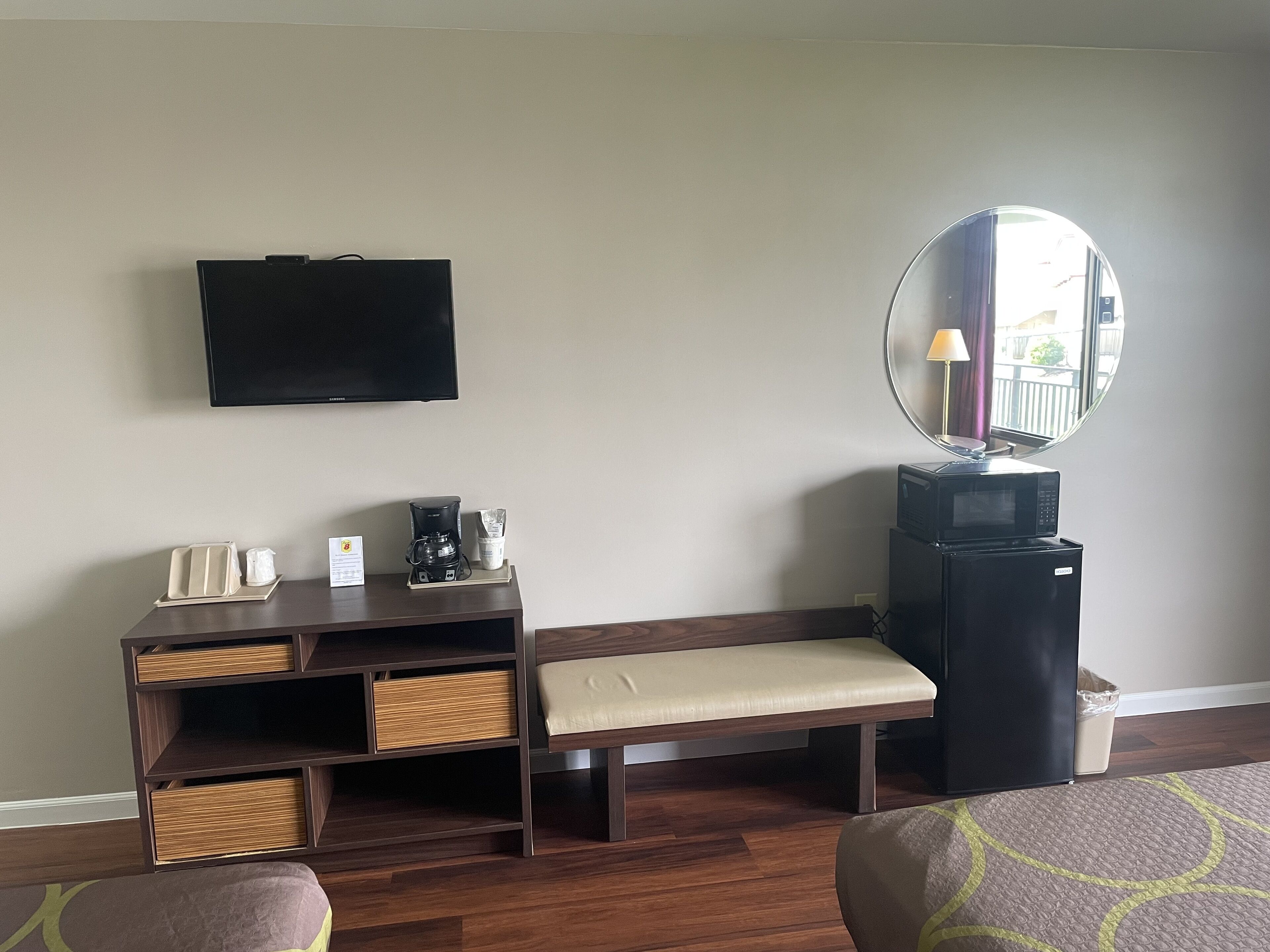 A hotel room with a flat-screen TV, a coffee maker, a bench, a mirror, and a trash bin.