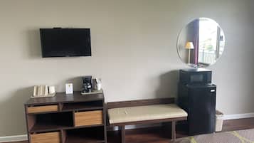 A hotel room with a flat-screen TV, a coffee maker, a bench, a mirror, and a trash bin.