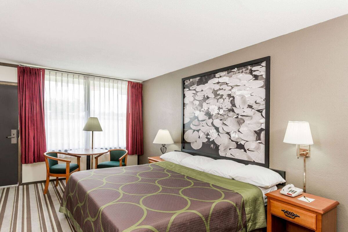 A hotel room with a bed, two chairs, a table, a lamp, a phone, and a large floral artwork on the wall.