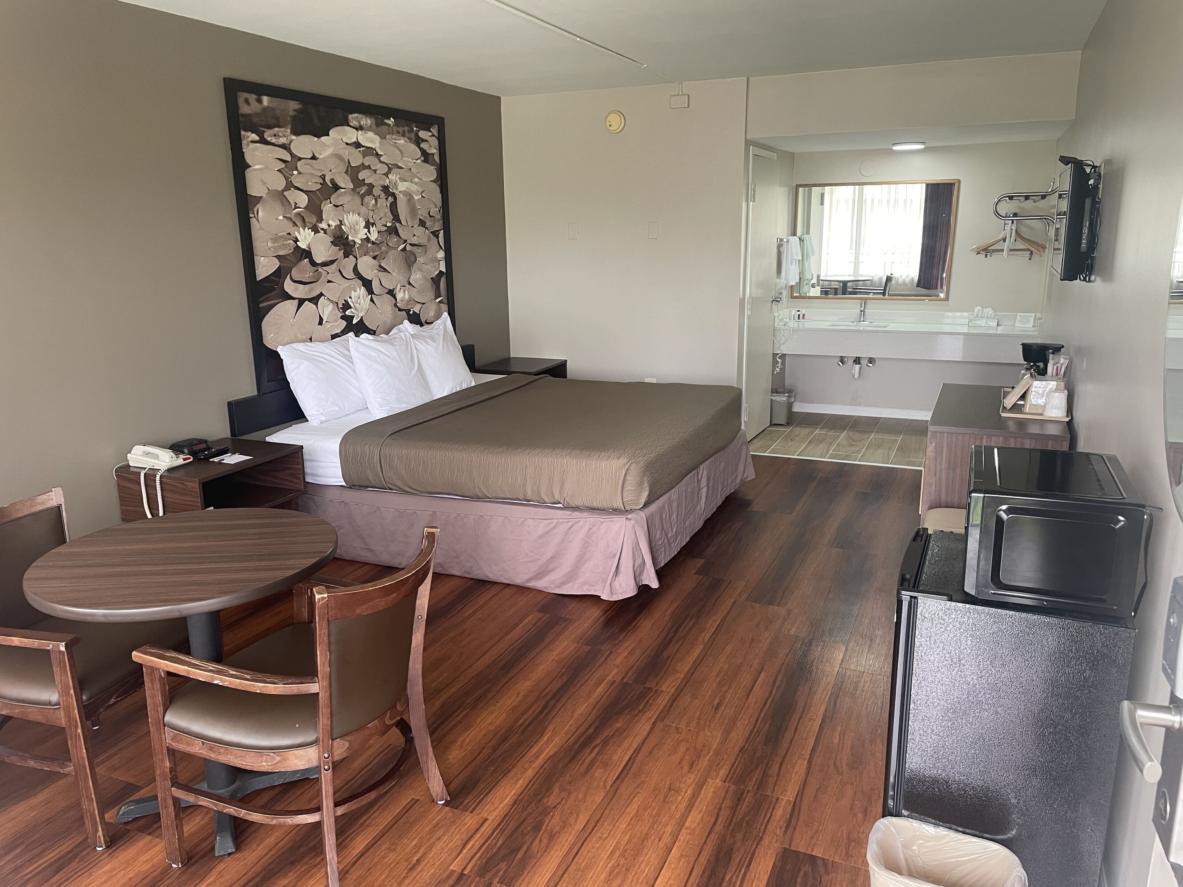 A hotel room with a bed, a desk with a chair, a small table, a TV, and a bathroom visible through an open door.
