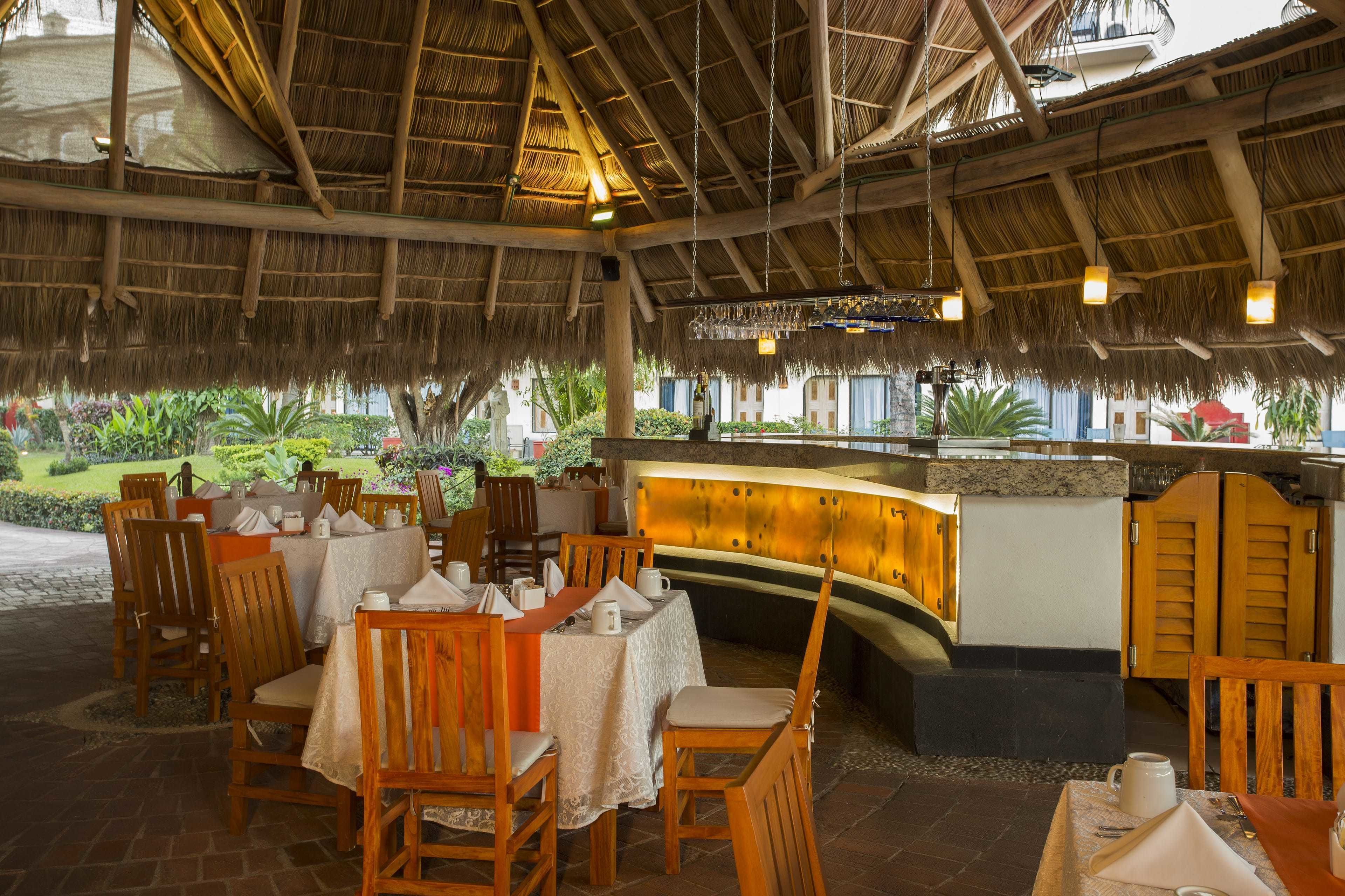 Un restaurant en plein air avec des tables et des chaises en bois, un toit de chaume et une vue sur la verdure.