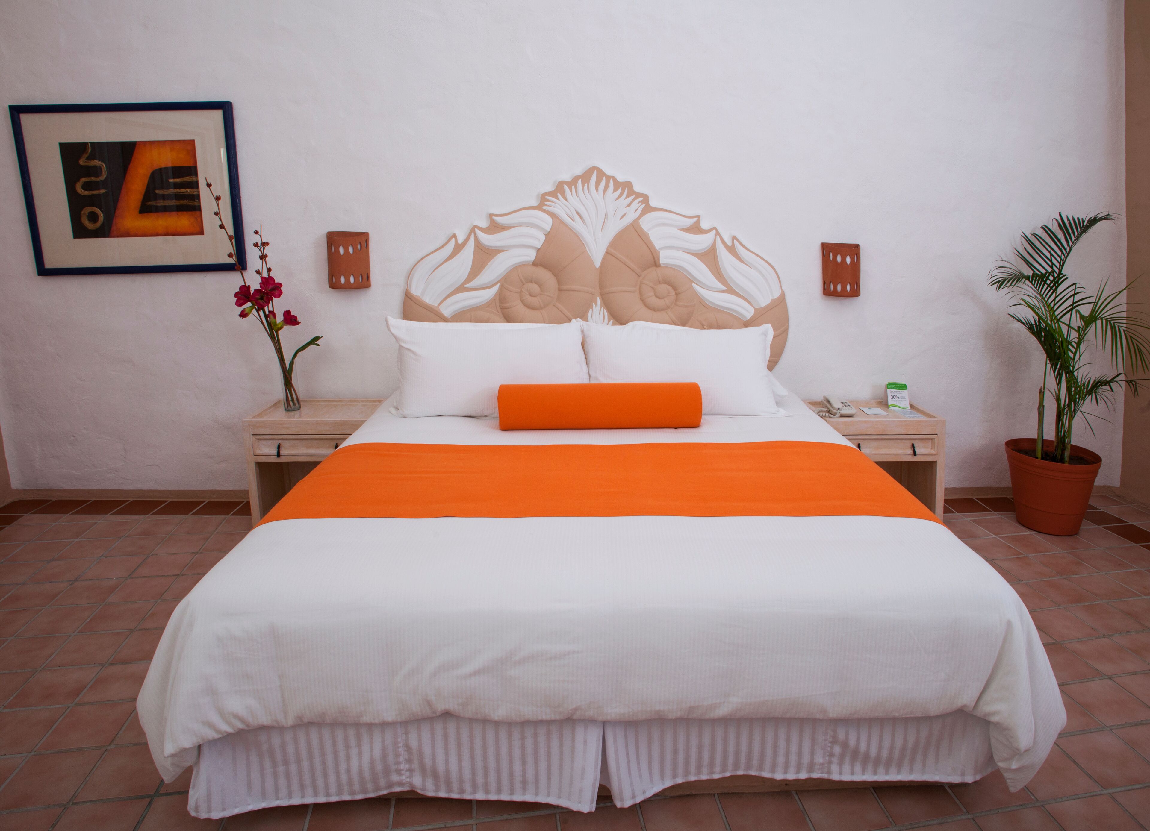 Een netjes opgemaakt bed met een oranje sierkussen, een decoratief hoofdbord en een ingelijst kunstwerk aan de muur.