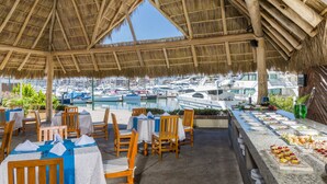 An outdoor dining area with wooden chairs and tables, a buffet with various food items, and a view of boats in the marina. - Flamingo Vallarta Hotel & Marina (Puerto Vallarta)