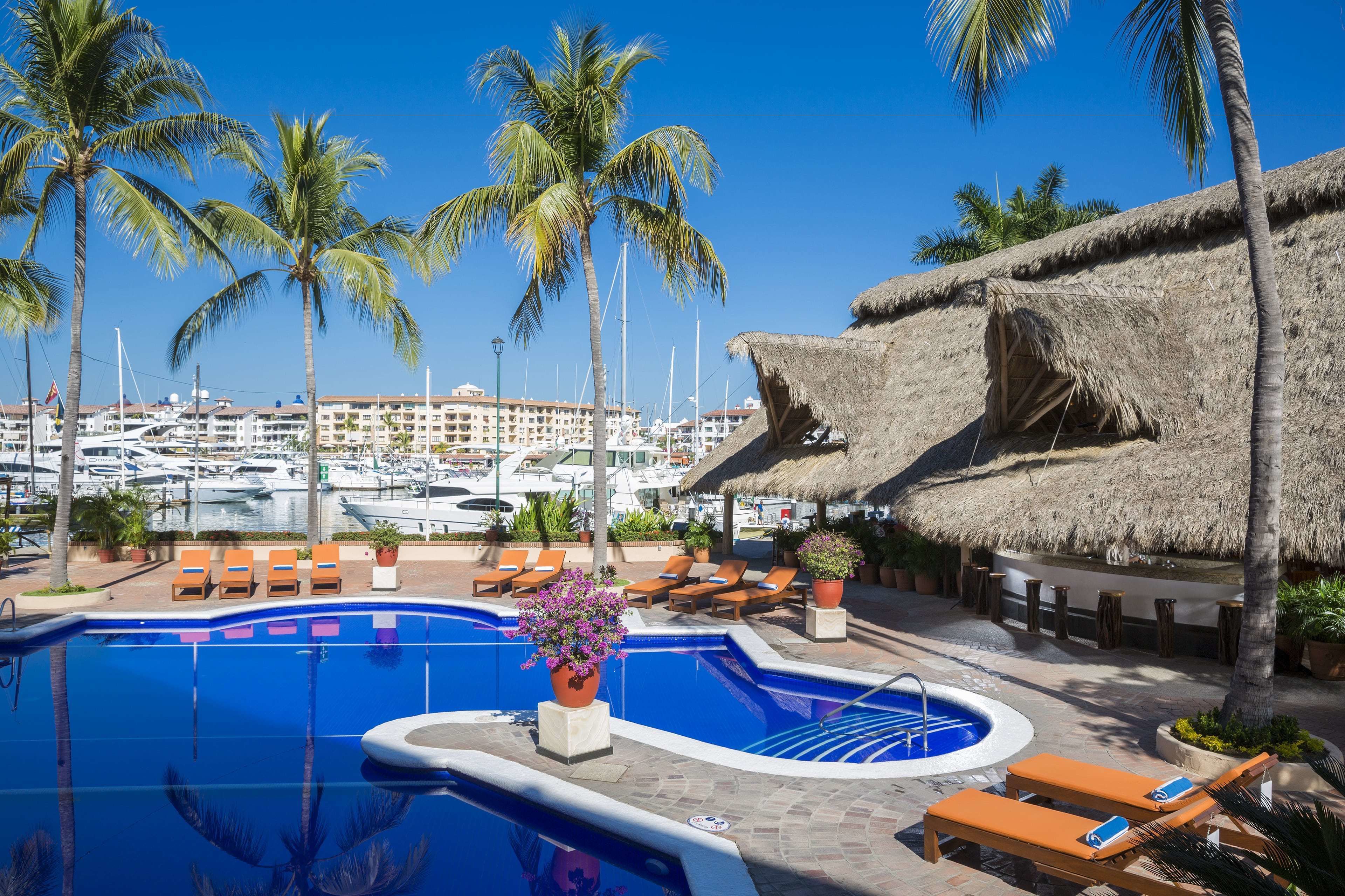 Zona de piscina con sillas de descanso, una estructura con techo de paja y vista a un puerto deportivo con barcos.