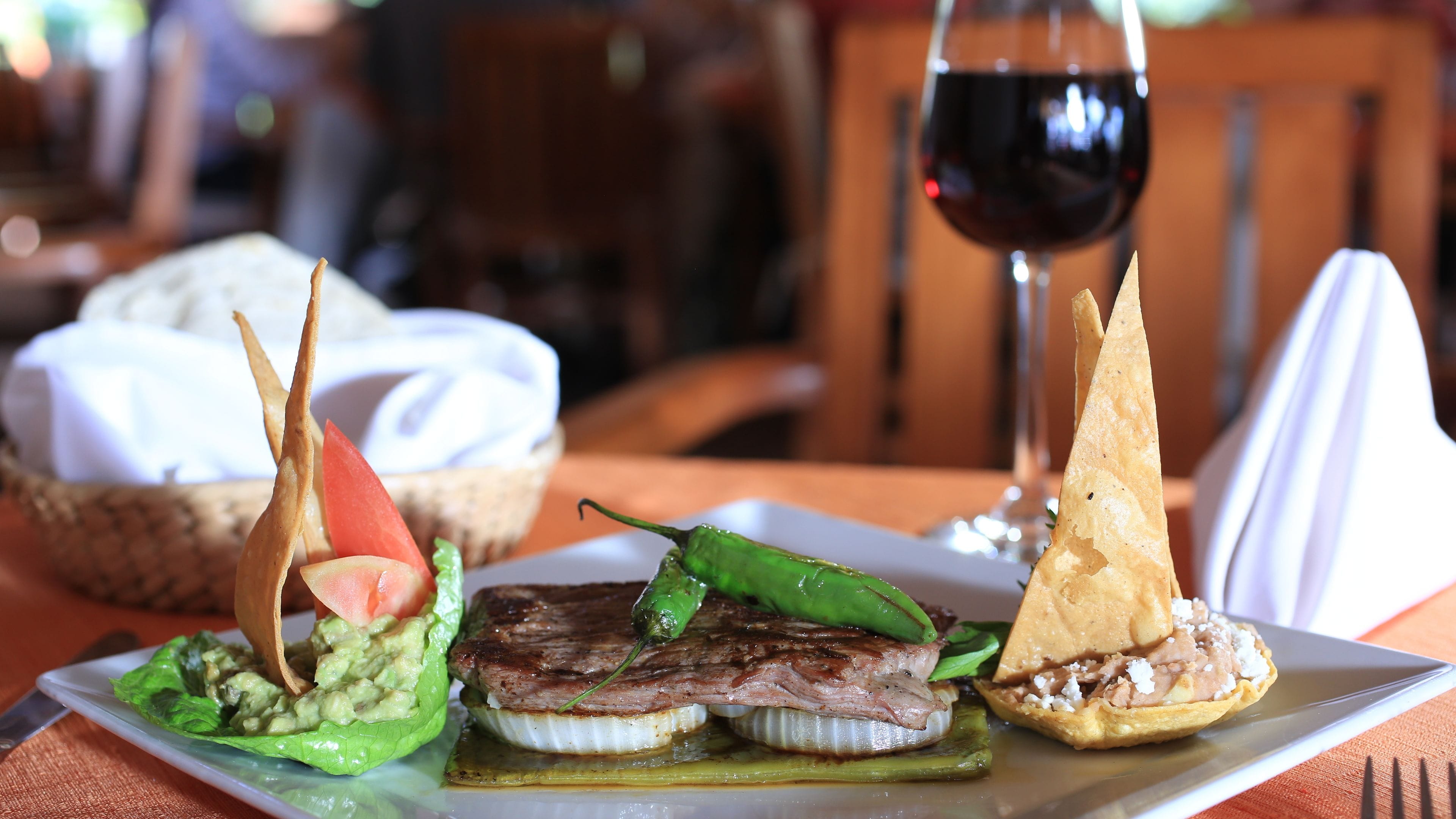 Une assiette garnie d’un steak, d’un avocat et d’un piment vert, accompagnée d’un verre de vin rouge.