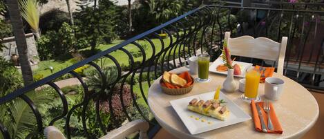 A balcony with a table set for breakfast, including toast, eggs, and juice, overlooking a lush garden.
