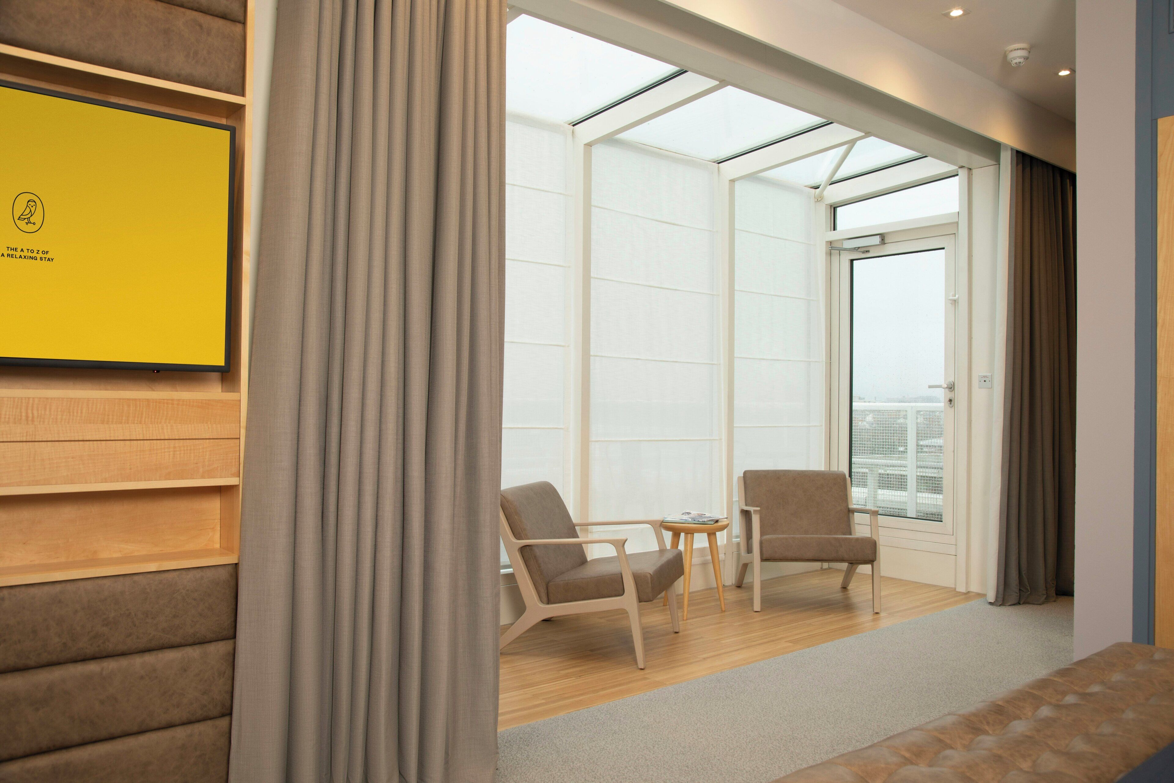A modern hotel room with a large window, two armchairs, a small table, and a yellow sign on the wall.
