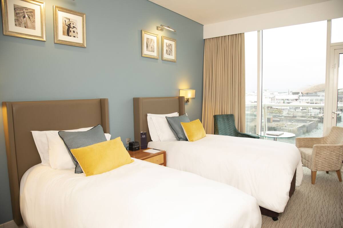 Habitación de hotel con dos camas, una mesita, una silla y un ventanal con vista a la ciudad.