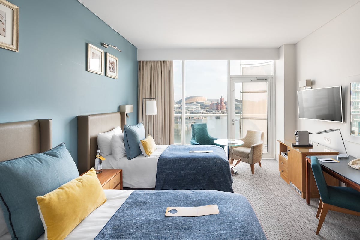 A hotel room with two beds, a desk, a chair, a TV, and a view of a cityscape.