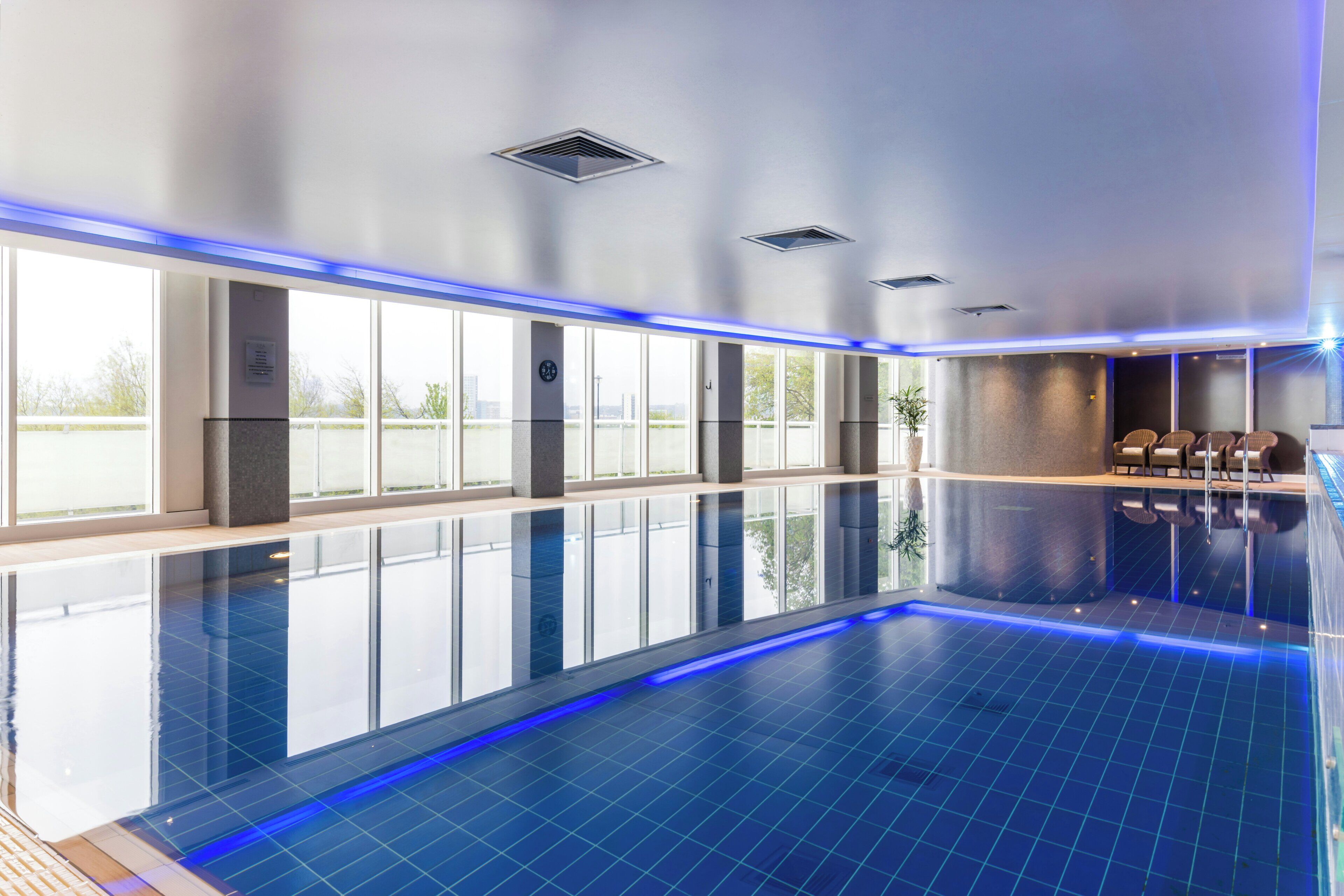 A large indoor swimming pool with clear blue water, surrounded by large windows offering views of the cityscape.