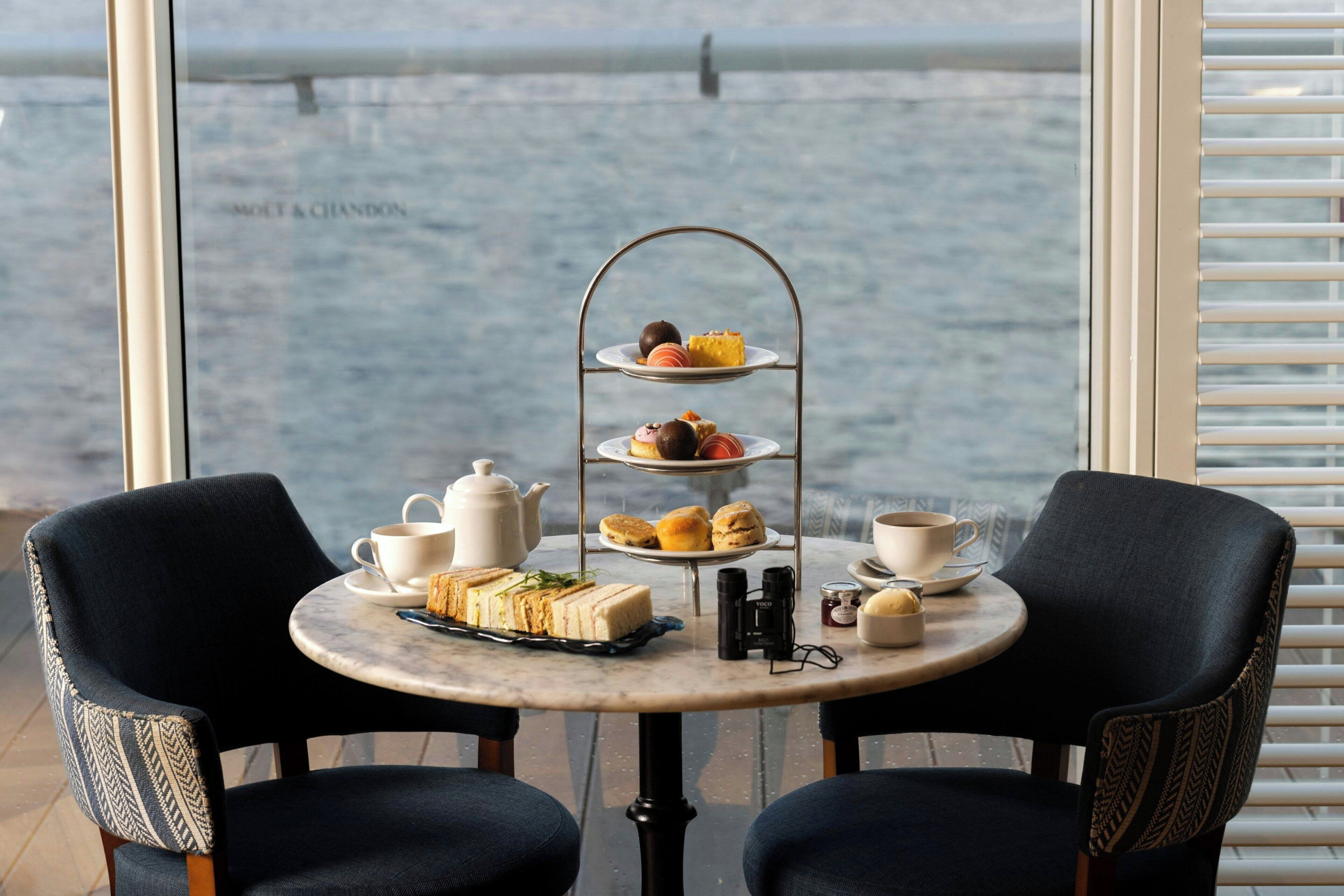 A table with a tiered dessert stand, a teapot, cups, and sandwiches.