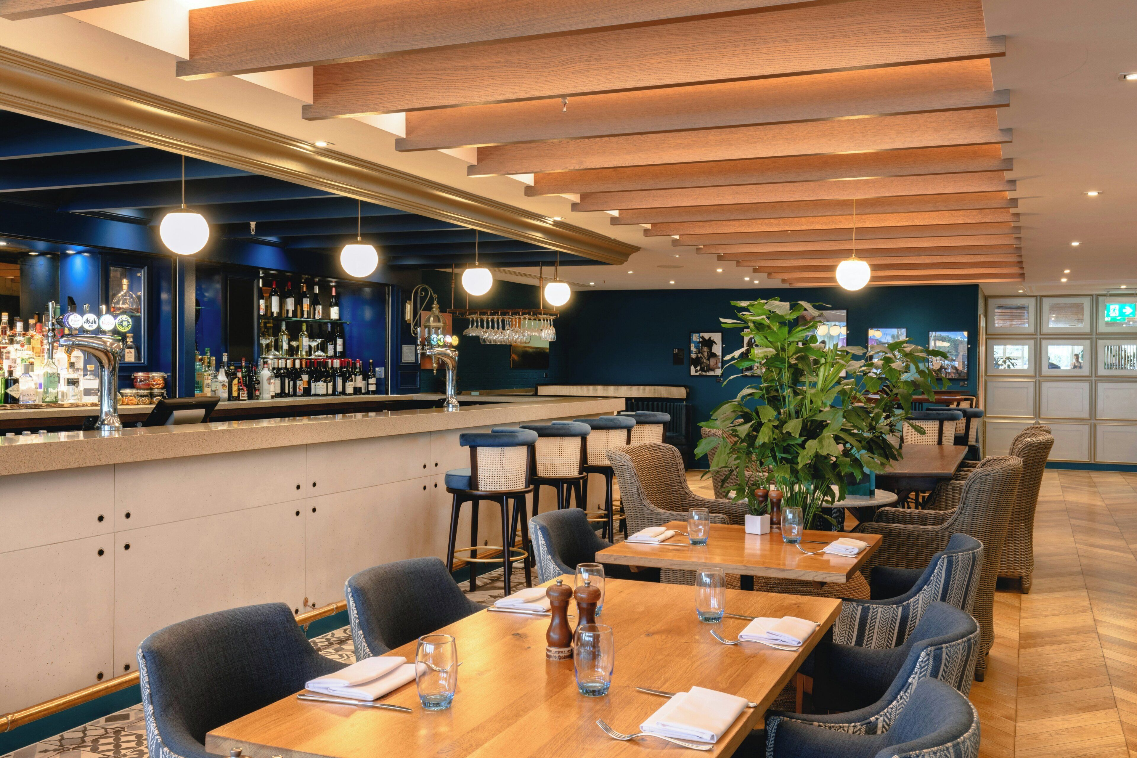 Eine Bar mit mehreren Zapfhähnen und Flaschenregalen, ein Sitzbereich mit Tischen und Stühlen sowie eine große Küsterpflanze.