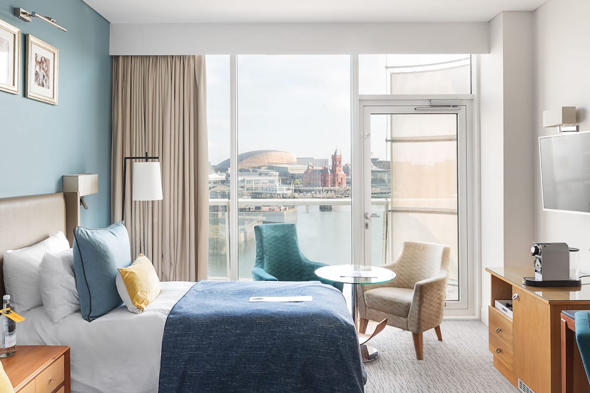 A hotel room with a bed, a desk, a chair, a TV, and a view of a cityscape.