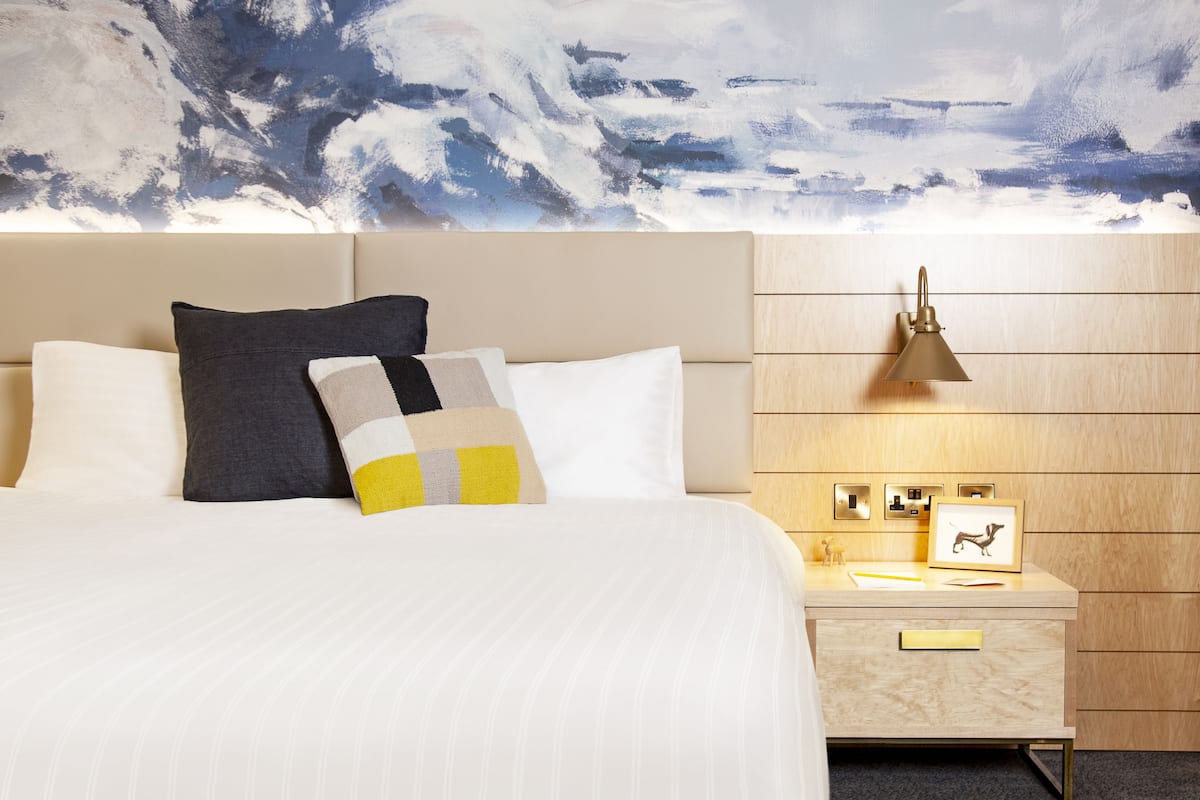 Una habitación de hotel con cama, almohadas, mesita de noche, lámpara y un cuadro enmarcado.