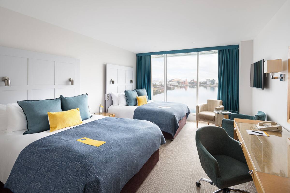 Habitación de hotel con dos camas, un escritorio, una silla y un ventanal con vistas a la ciudad.