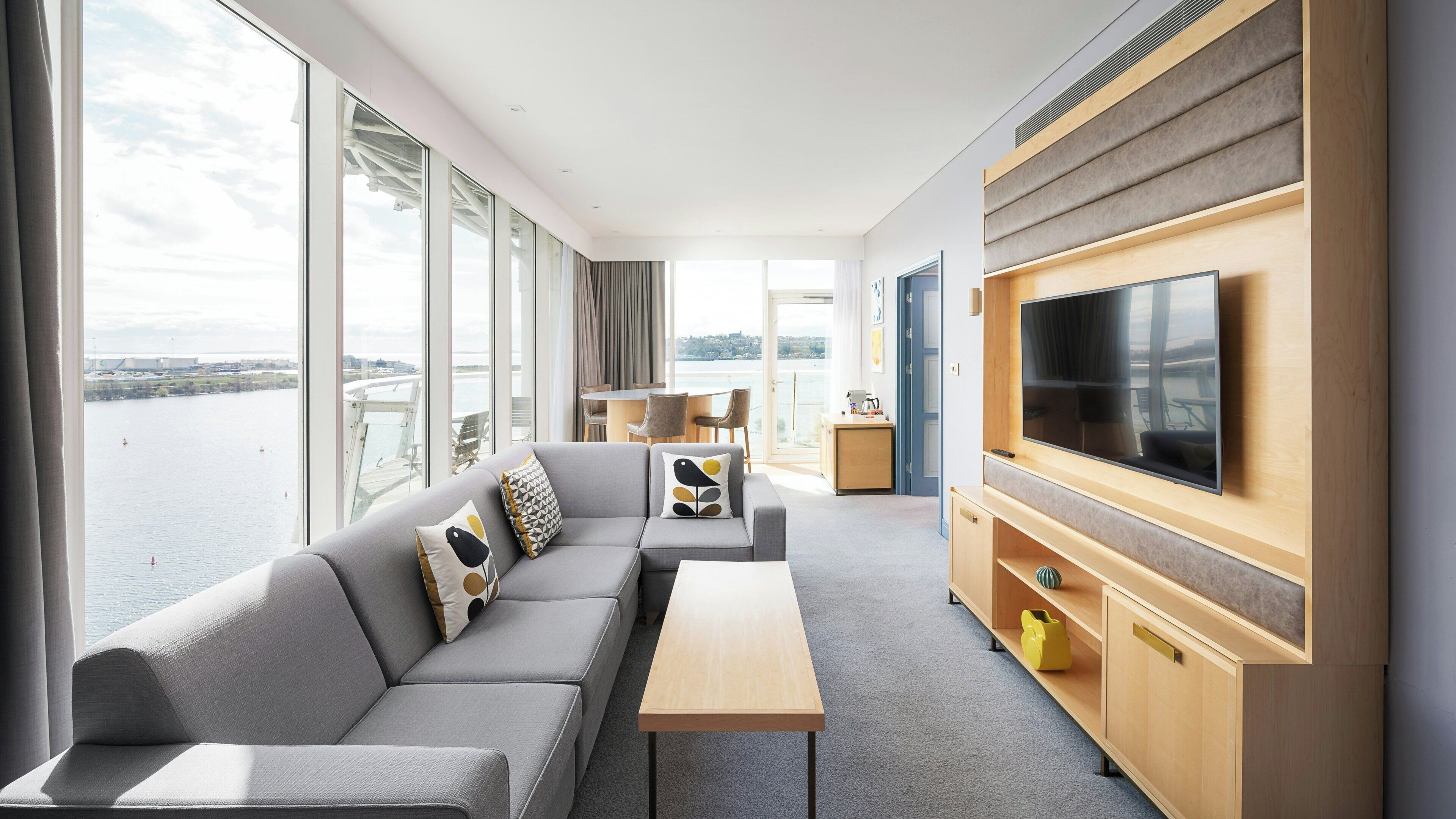 A modern hotel room with a sofa, a TV, a table, and a view of the water.