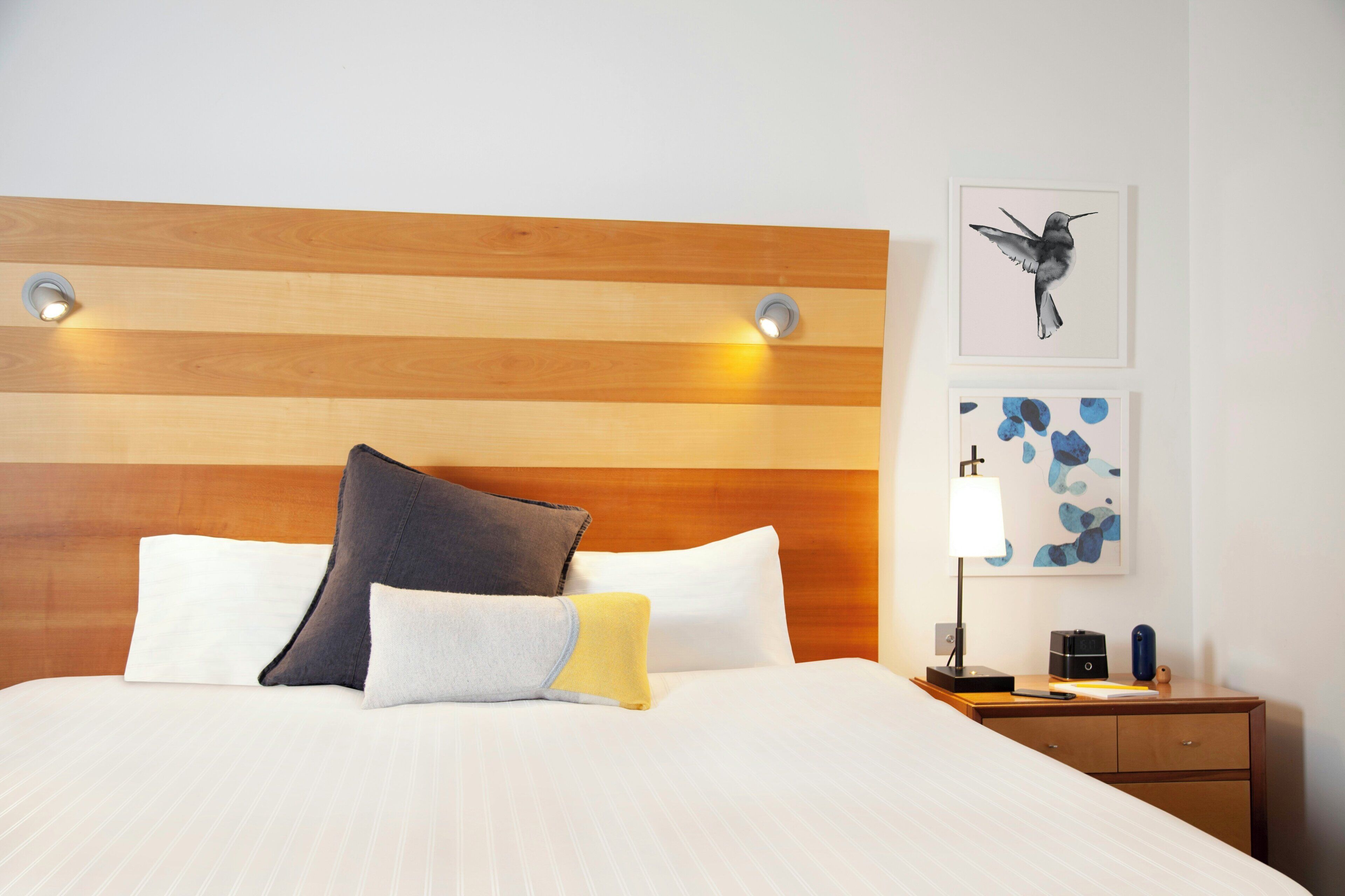 A hotel room with a wooden headboard, a bed with multiple pillows, a nightstand with a lamp, and artwork on the wall.