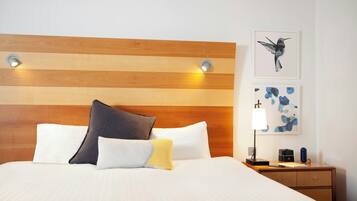 A hotel room with a wooden headboard, a bed with multiple pillows, a nightstand with a lamp, and artwork on the wall.