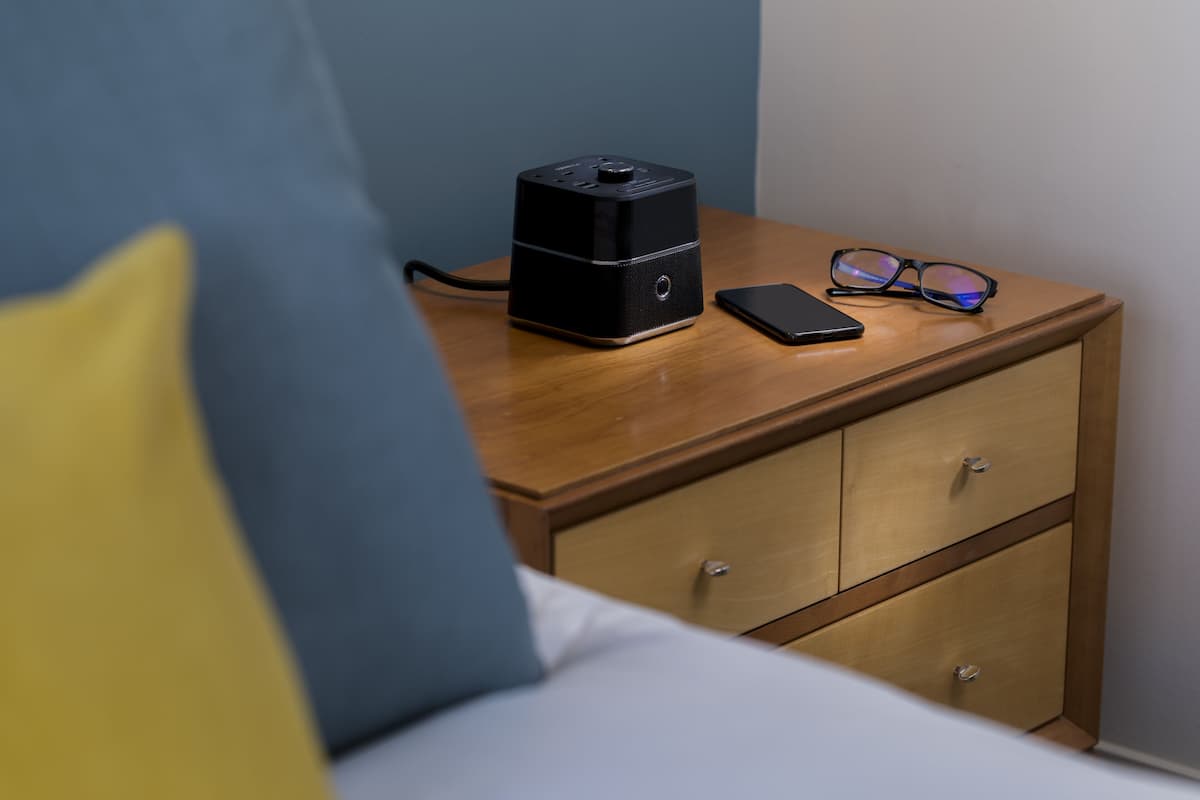 A wooden bedside table with a black electronic device, a smartphone, and a pair of glasses.