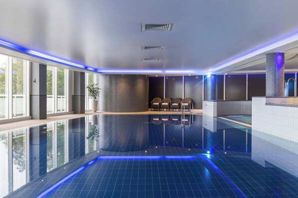 A modern indoor swimming pool with clear blue tiles, a seating area with chairs, and a large window providing natural light.