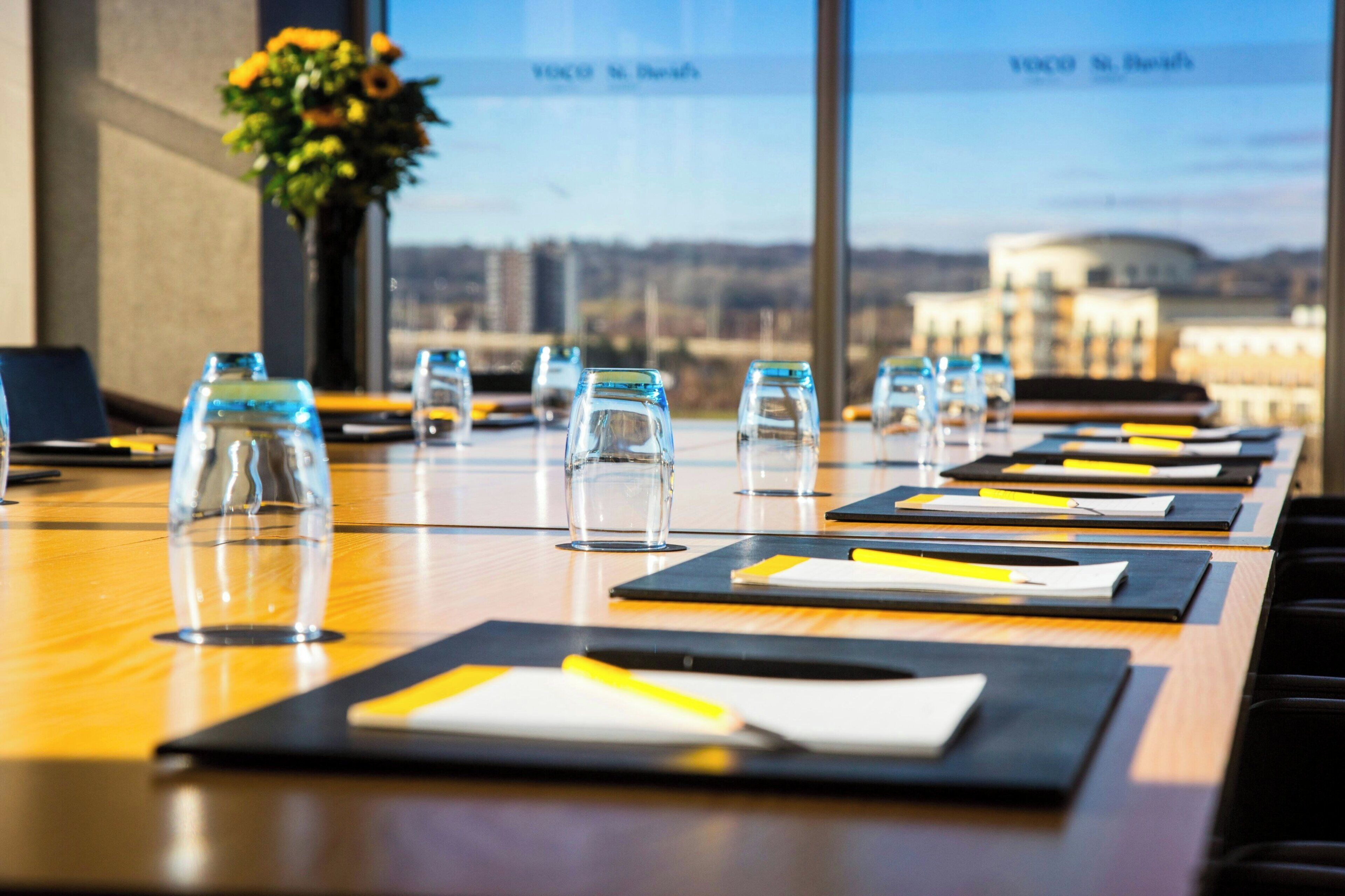 A conference table with documents, pens, and glasses, set against a backdrop of a cityscape through large windows.