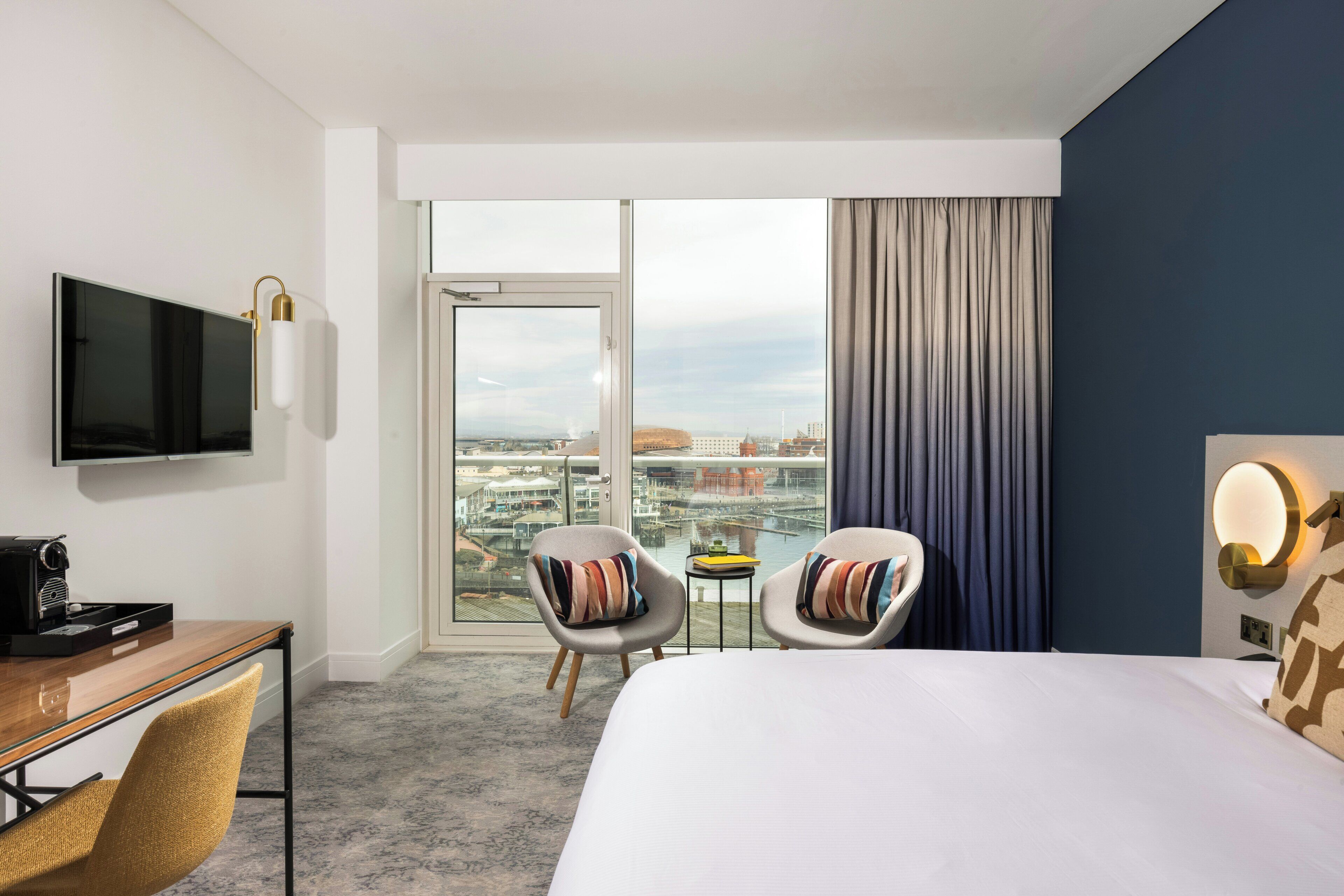 Une chambre d’hôtel comprenant un lit, deux fauteuils, un bureau avec une machine à café, une télévision et une vue sur le paysage urbain par une grande fenêtre.