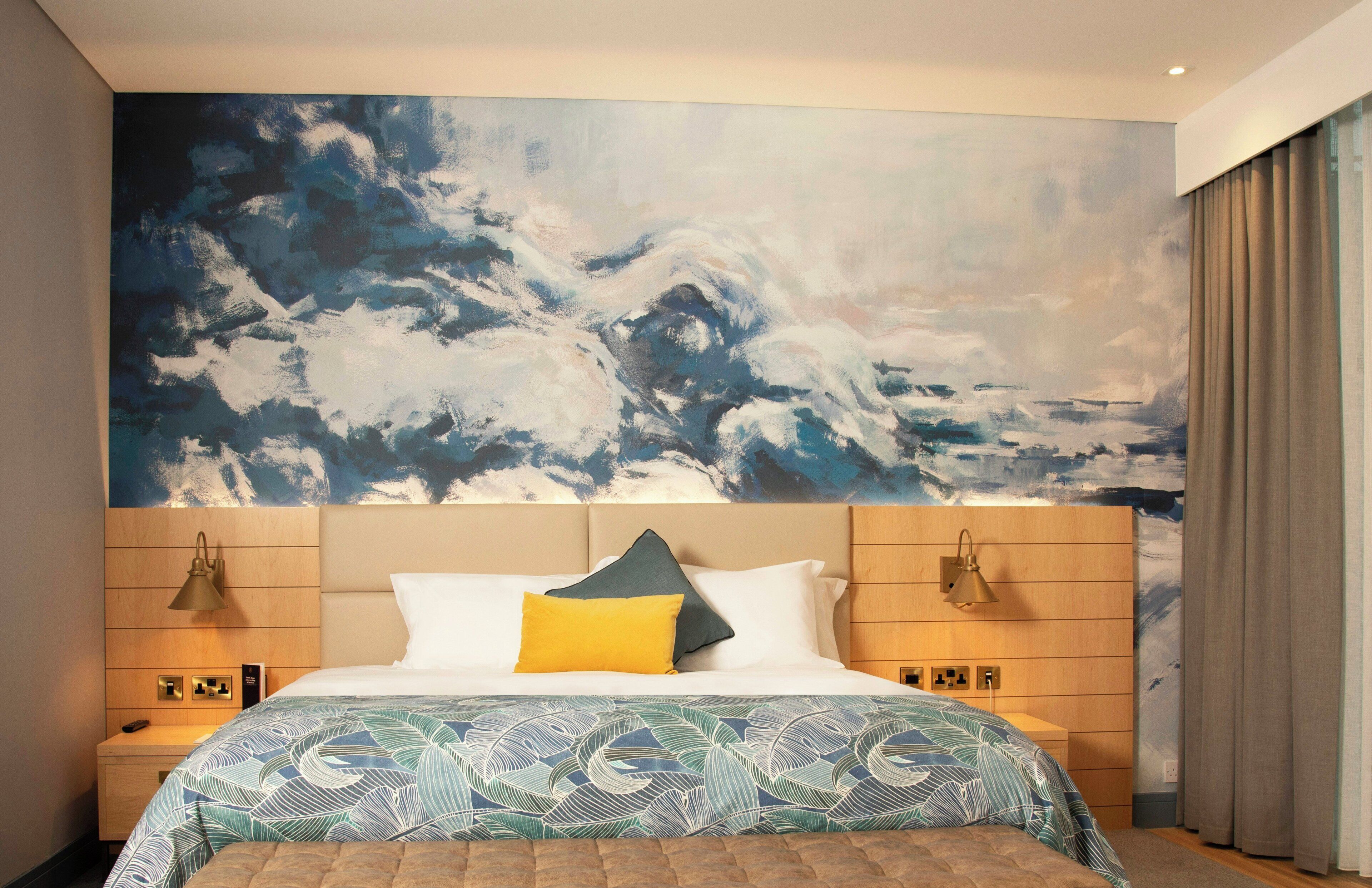A hotel room with a large abstract painting on the wall, a bed with a patterned bedspread, two bedside lamps, and a wooden headboard.