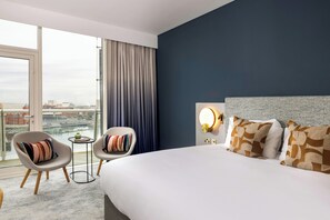 A hotel room with a large bed, two armchairs, a small table, and a view of a cityscape through a large window. - voco St David's Cardiff by IHG (Cardiff)