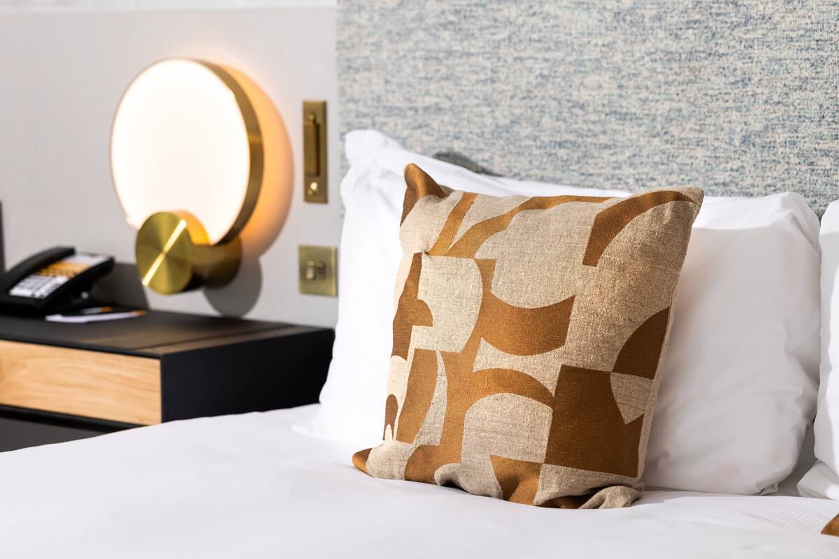 Una cama bien tendida con un almohadón estampado, una mesita de noche con una lámpara y una pared con textura.