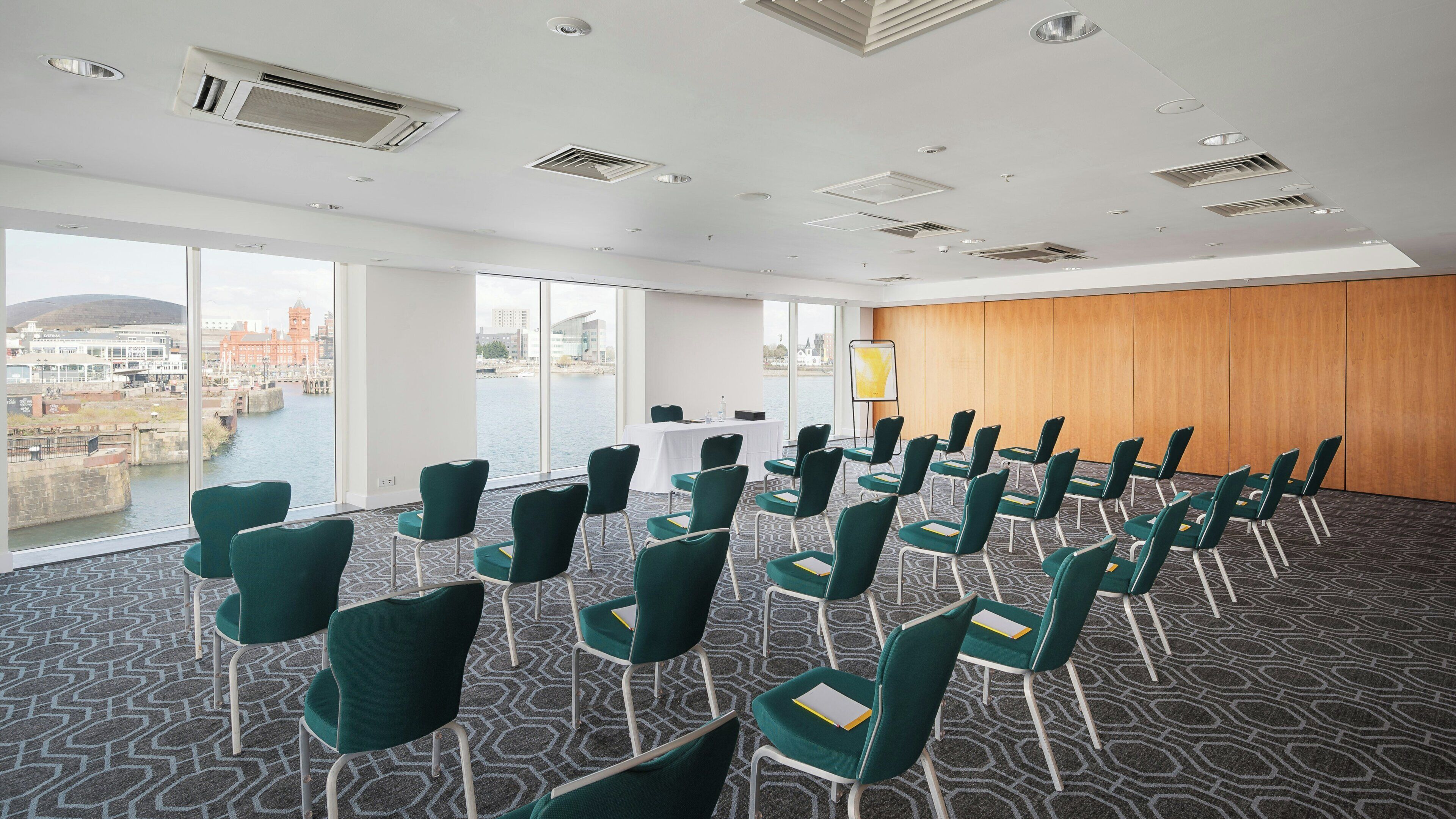 A conference room with a view of a waterfront, equipped with green chairs, a white table, and a presentation screen.