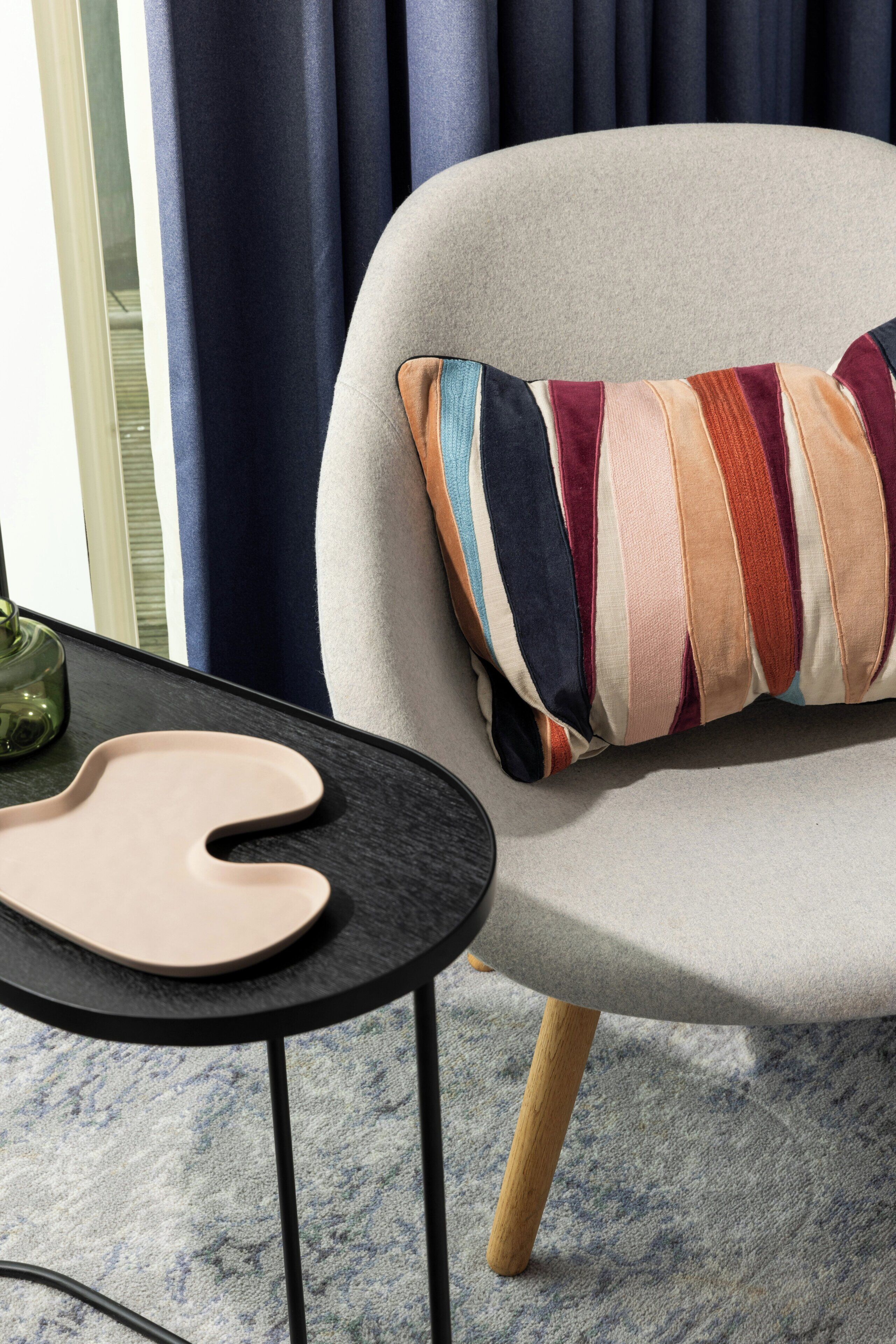 A modern chair with a multicolored striped pillow, a black side table with a beige object, and a window with dark curtains.