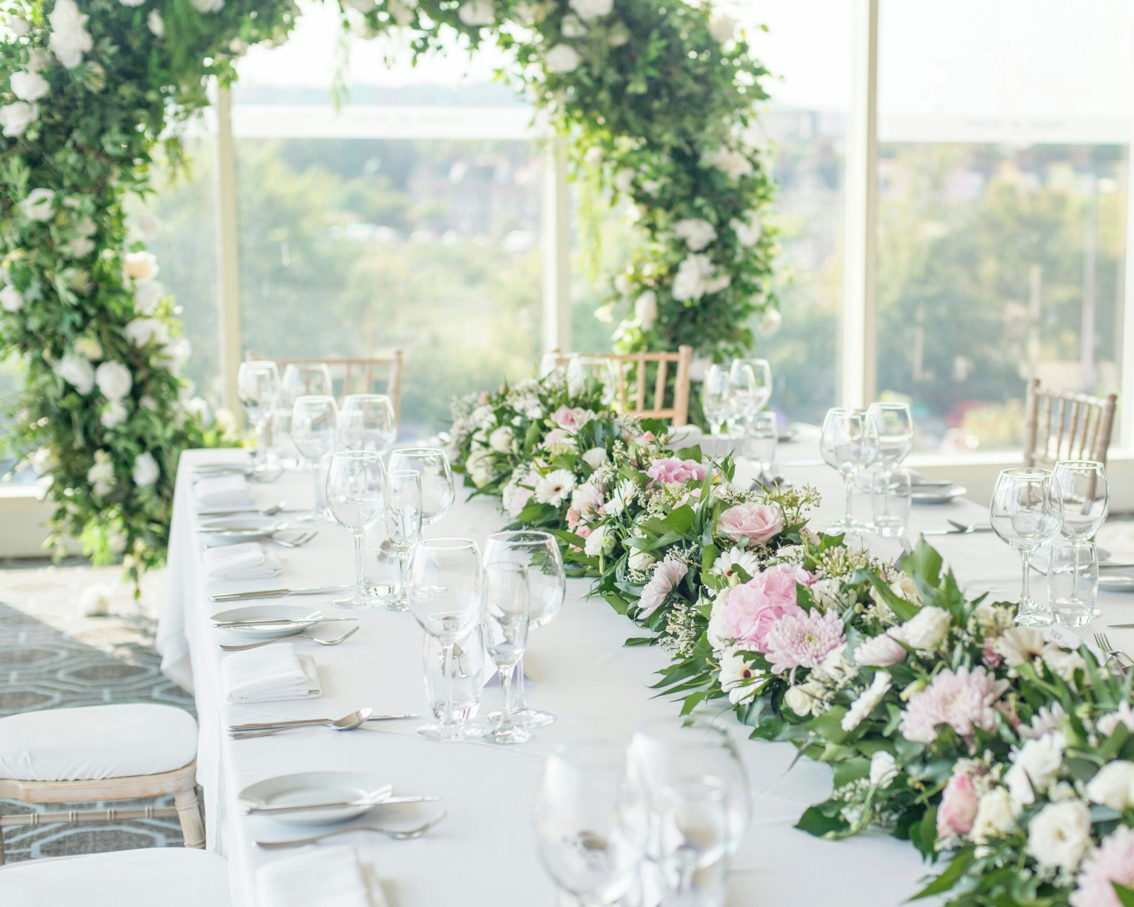 A table set for a formal event with clear glassware and white tablecloths, surrounded by floral arrangements.
