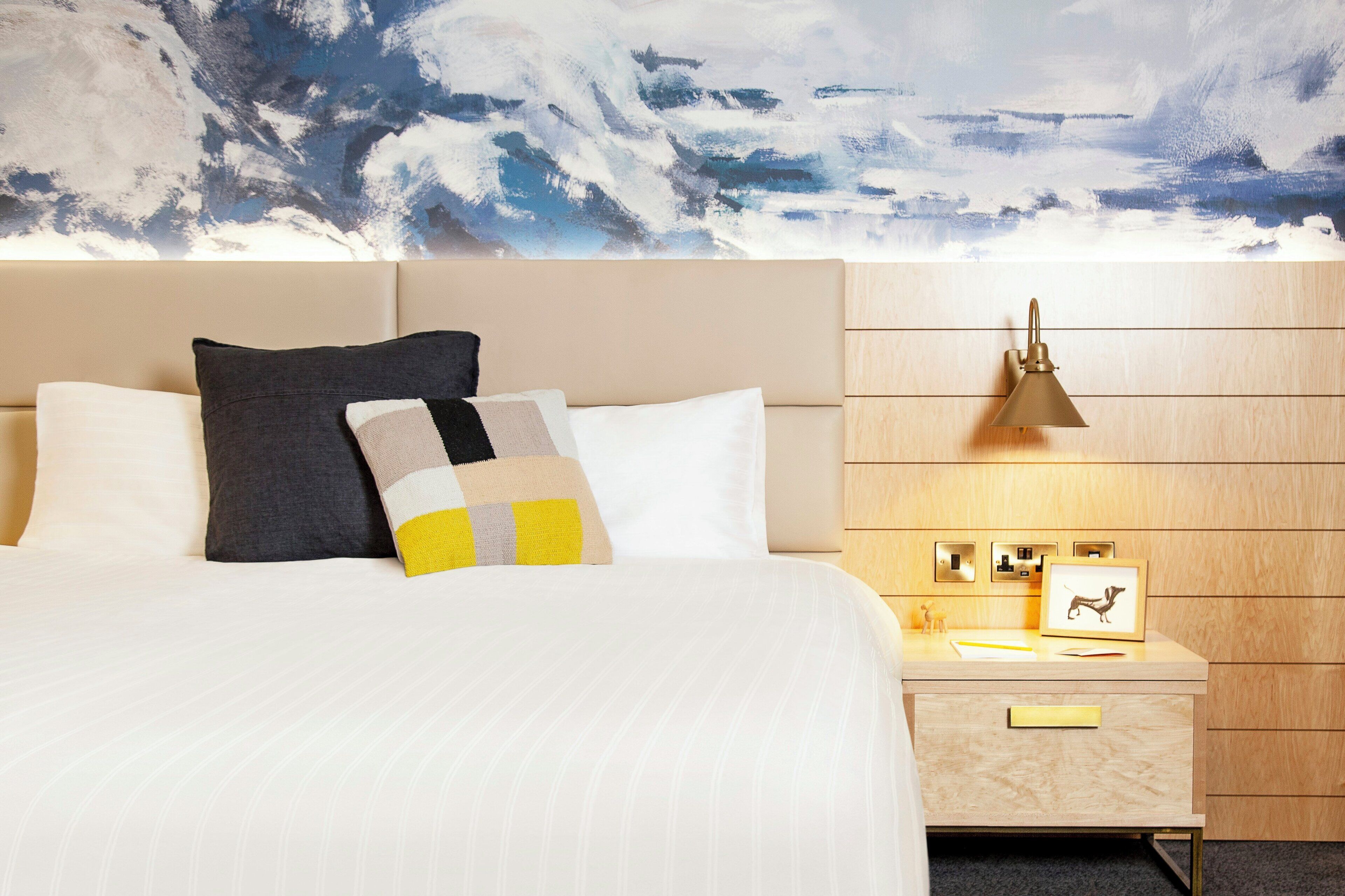 A hotel room with a bed, pillows, a nightstand, a lamp, and a wall-mounted picture.
