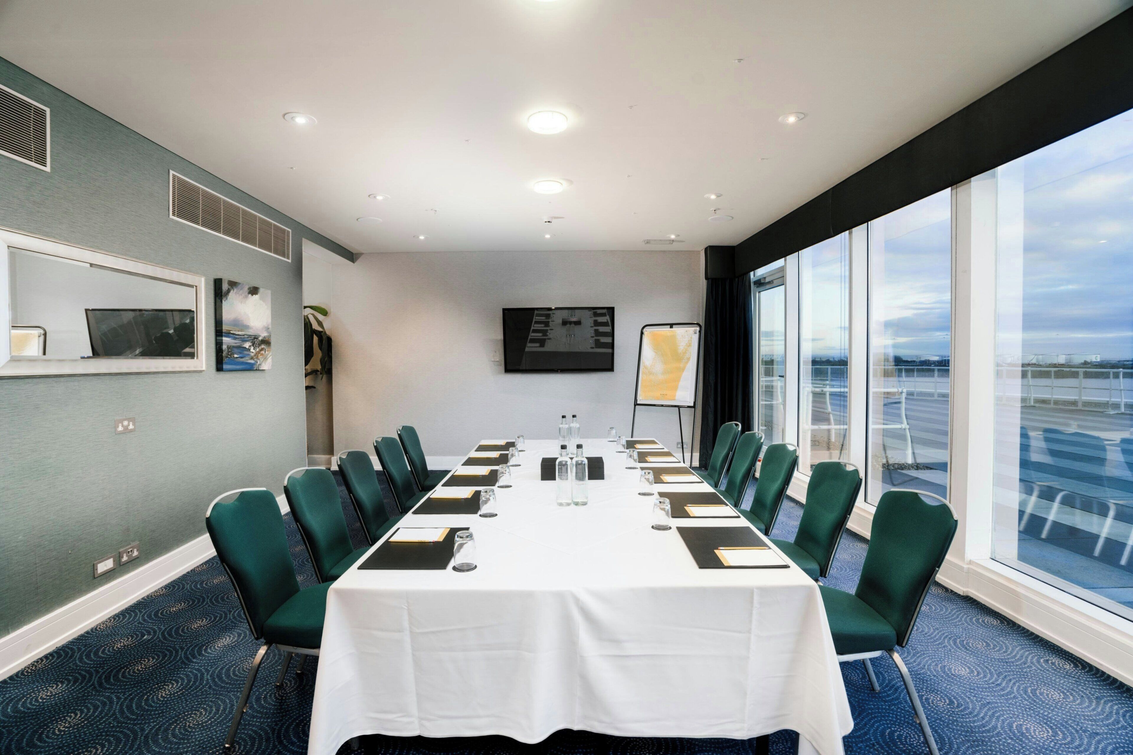 Un salón de conferencias con una larga mesa, sillas verdes, un mantel blanco, una pantalla de proyector y un ventanal con vistas.