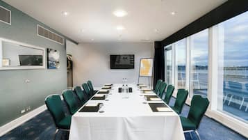 Un salón de conferencias con una larga mesa, sillas verdes, un mantel blanco, una pantalla de proyector y un ventanal con vistas.