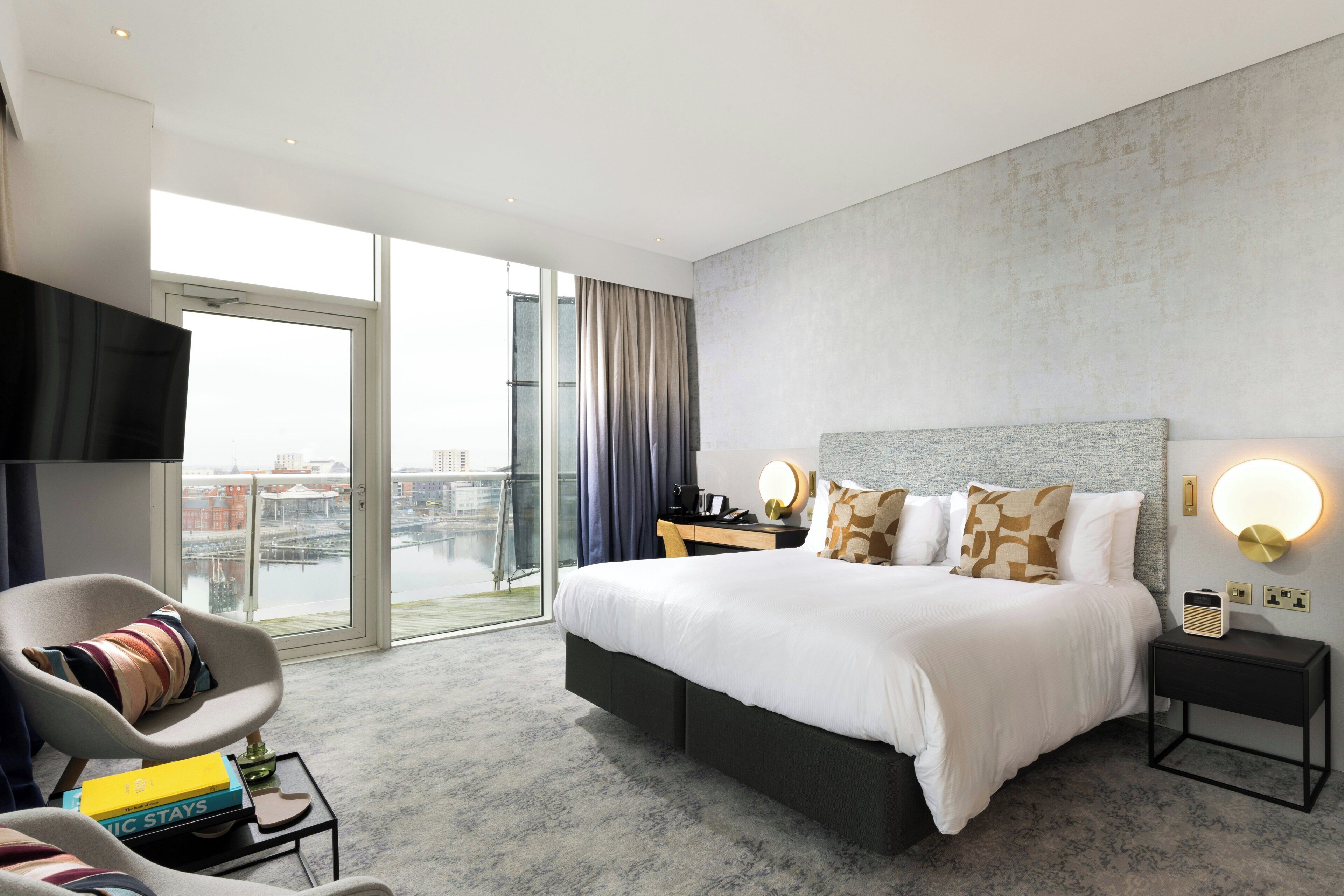A modern hotel room with a large bed, a seating area with a chair and books, a TV, and a view of the city through a large window.
