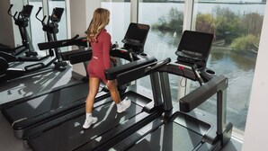 Une femme marche sur un tapis de course dans une salle de sport dotée de grandes fenêtres donnant sur un plan d’eau.