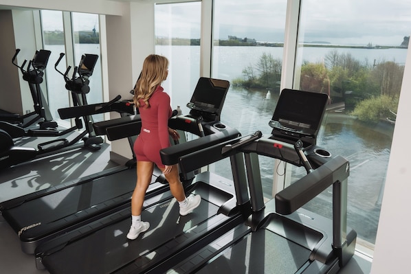 A woman walking on a treadmill in a gym with large windows overlooking a body of water. - voco St David's Cardiff by IHG (Cardiff)