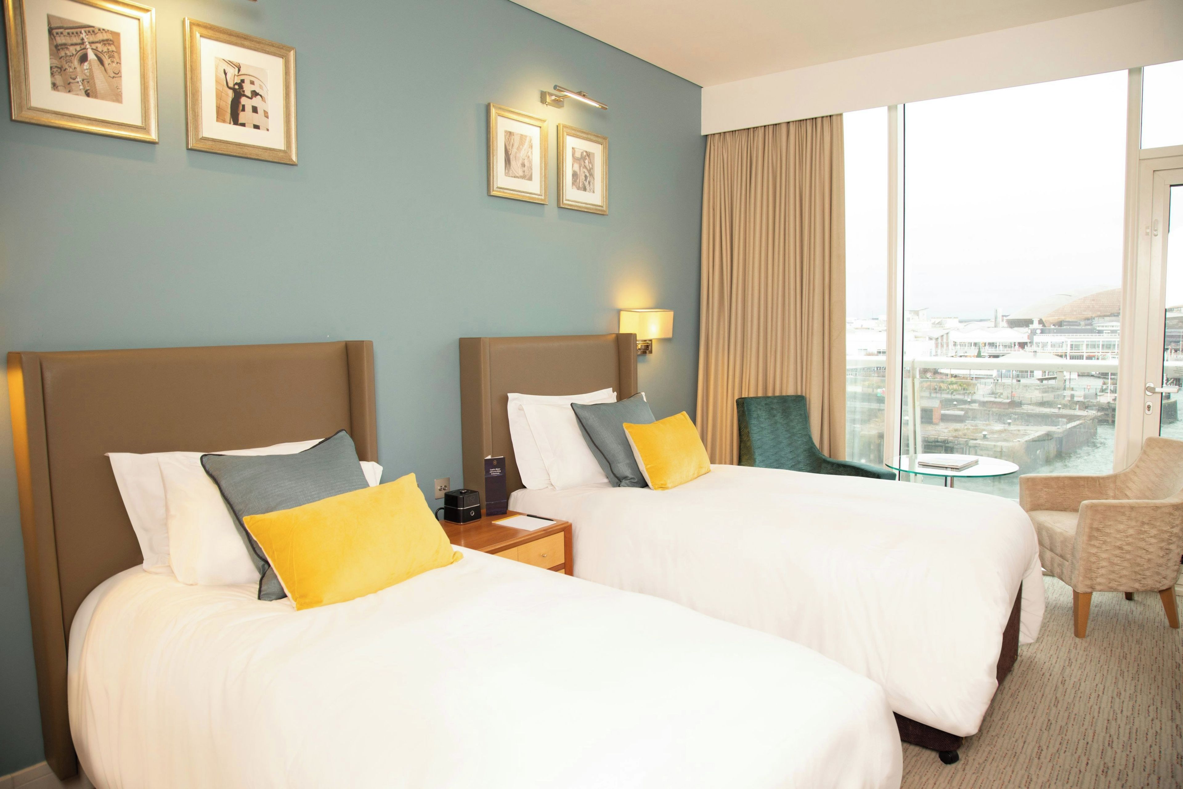 Habitación de hotel con dos camas, una mesita de noche, una silla y vistas a la ciudad.