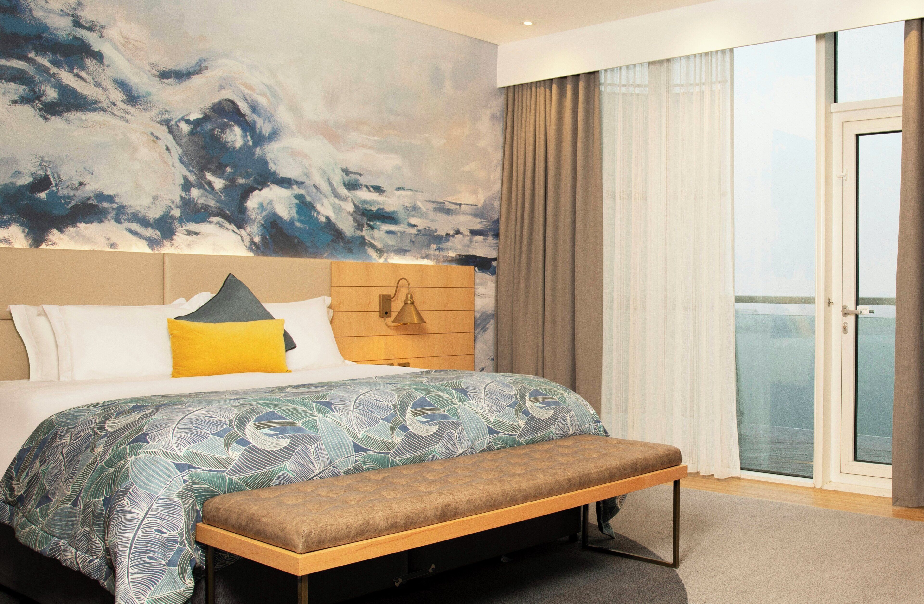A hotel room with a large bed, a bench, a headboard with a light fixture, a large abstract painting on the wall, and a view through the window.