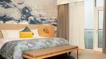 A hotel room with a large bed, a bench, a headboard with a light fixture, a large abstract painting on the wall, and a view through the window.