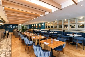A restaurant with wooden tables and blue upholstered chairs, a patterned floor, and a view of the city through large windows.