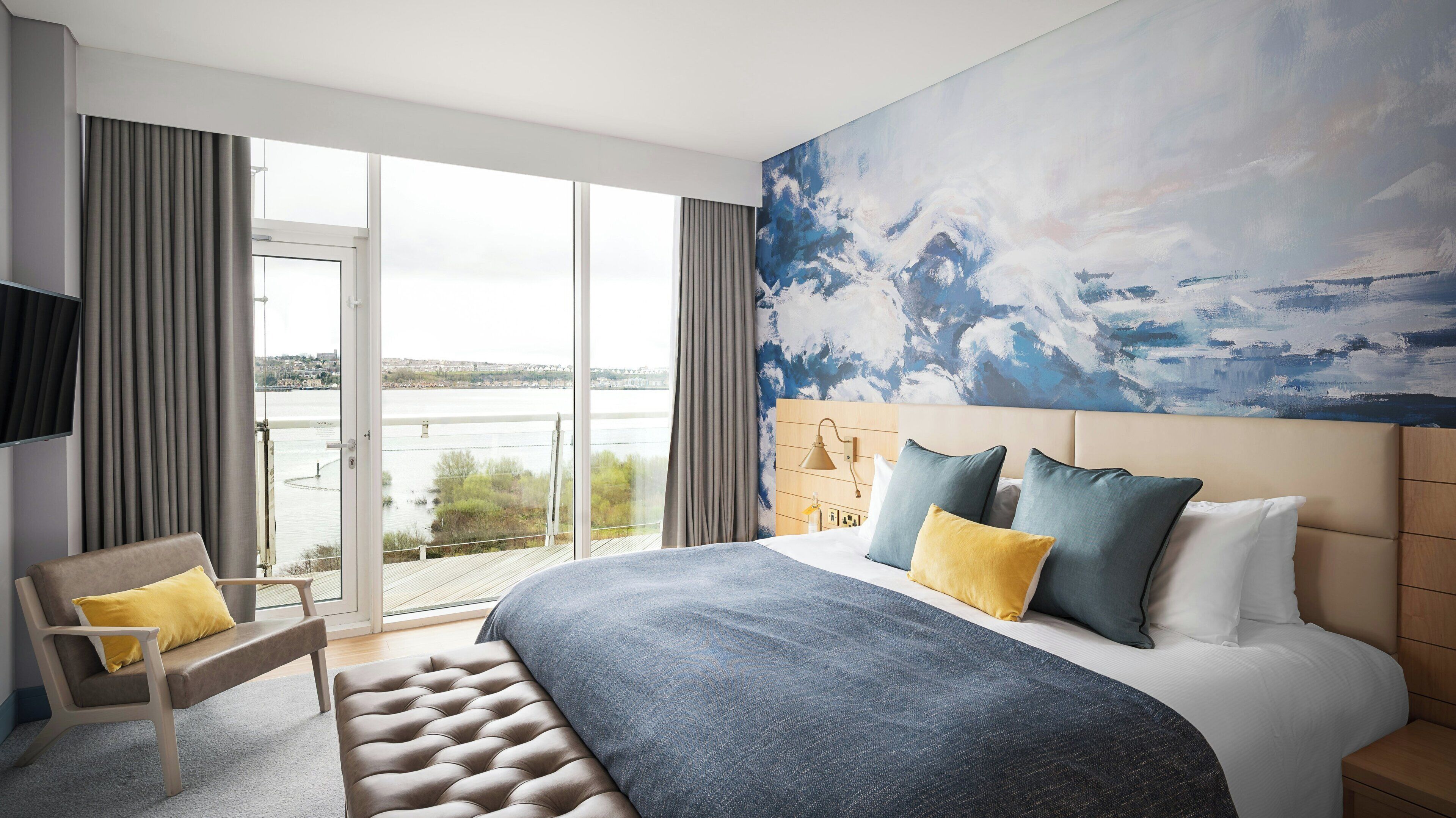 A hotel room with a large bed, a view of the outdoors, a painting on the wall, and a television on the left.