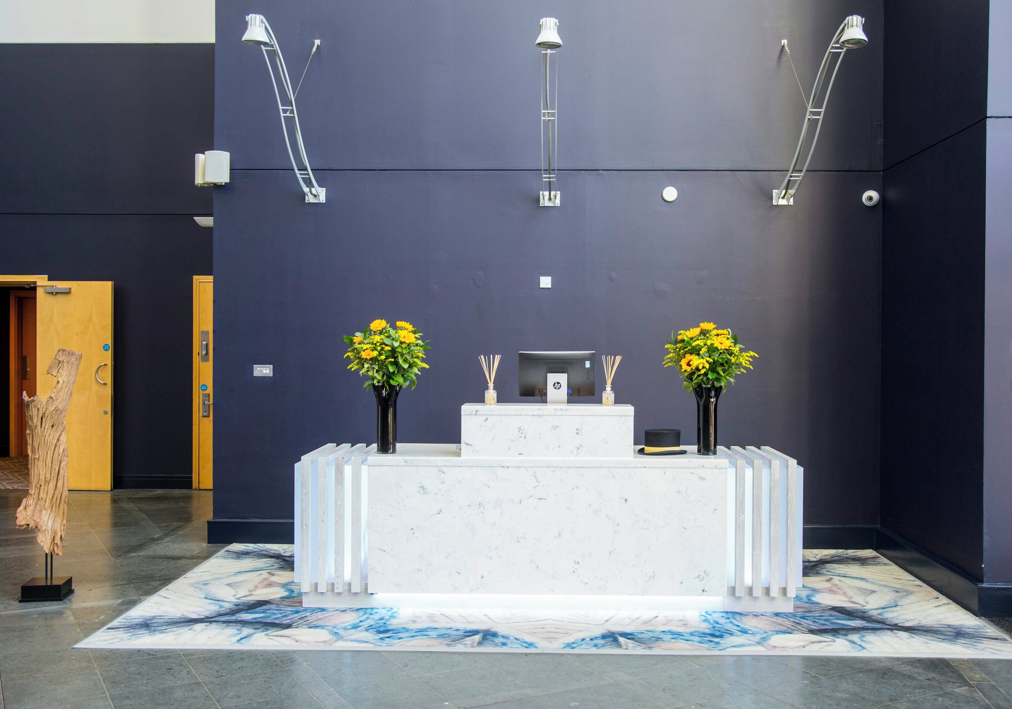 A reception desk with two flower arrangements, a marble floor, and a dark wall with ceiling lights.