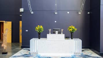 Un mostrador de recepción con dos arreglos florales, piso de mármol y pared oscura con luces de techo.