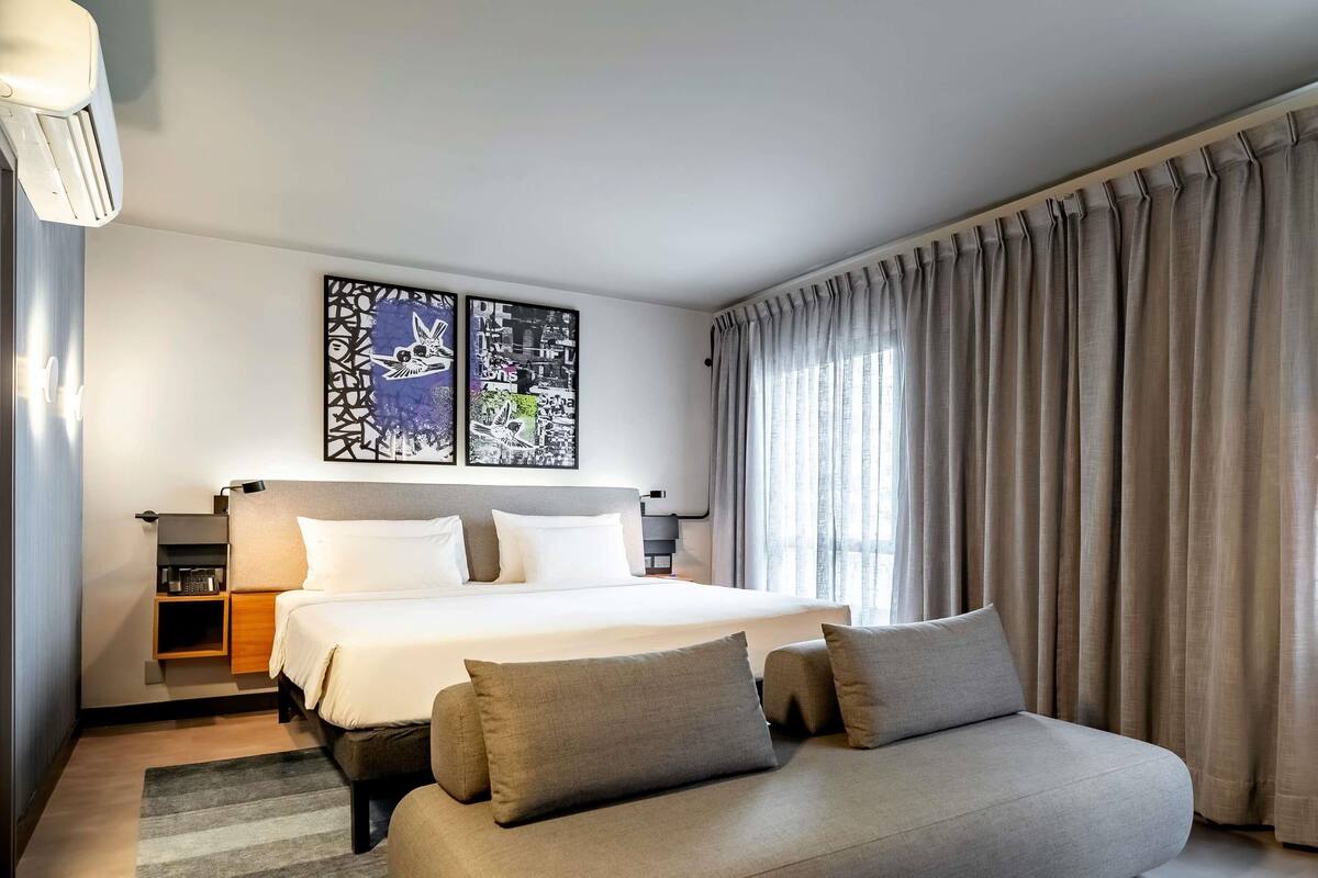 Ein modernes Hotelzimmer mit einem großen Bett, einer Couch, einem Nachttisch und zwei an der Wand hängenden Kunstwerken.