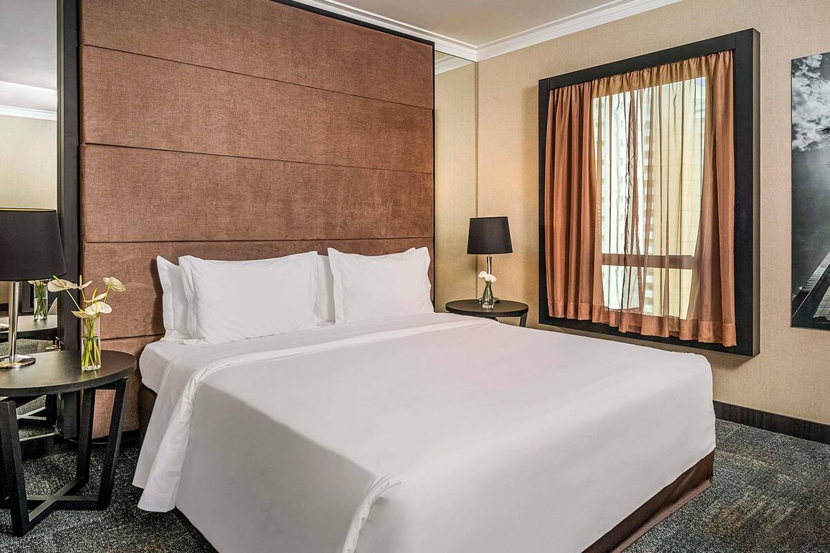 Ein Hotelzimmer mit einem großen Bett, einem Nachttisch mit Lampe, einem kleinen Tisch mit Blumenvase und einem Fenster mit Vorhängen.